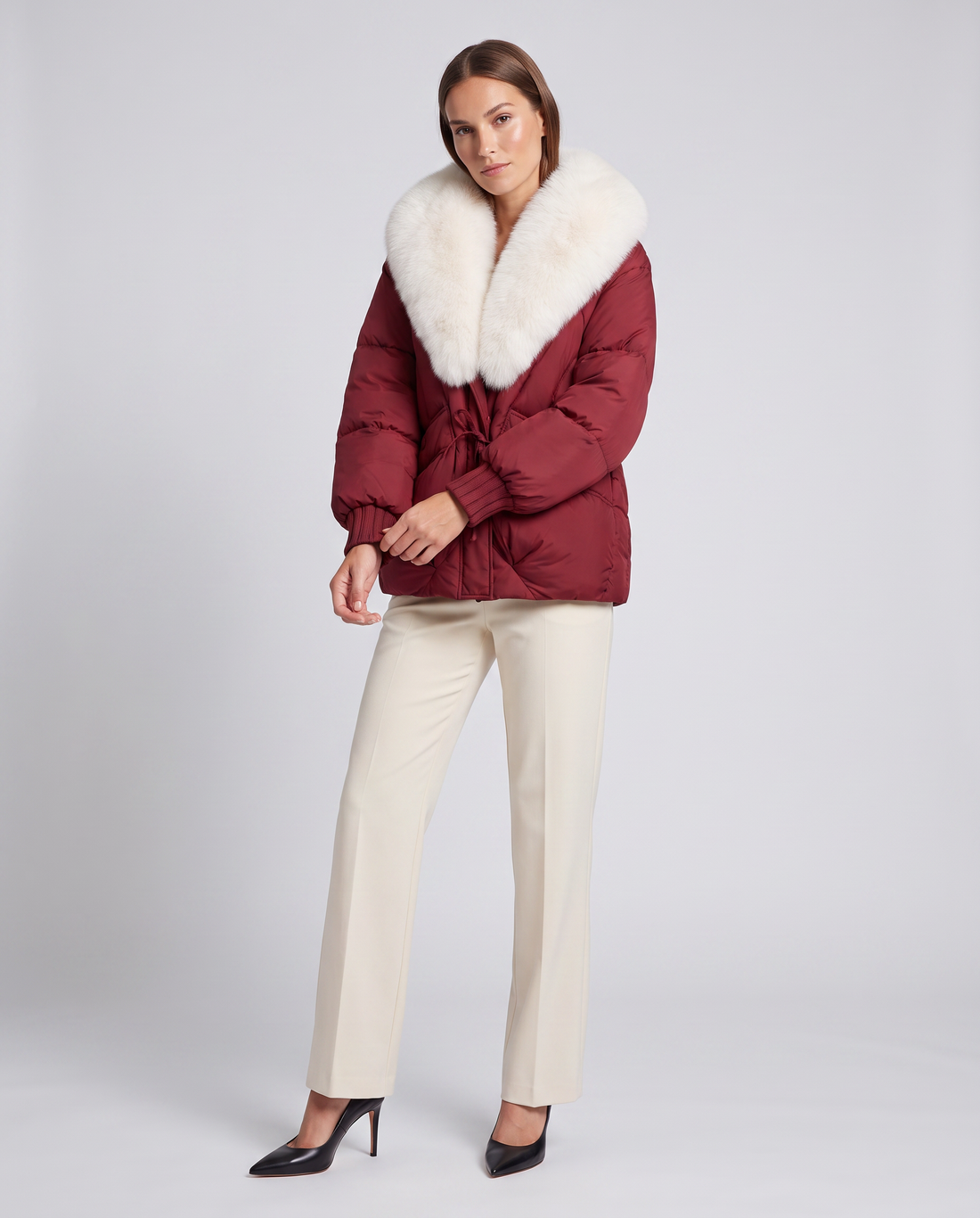 Woman wearing burgundy puffer jacket with white faux fur collar, cream pants, and black pointed-toe heels