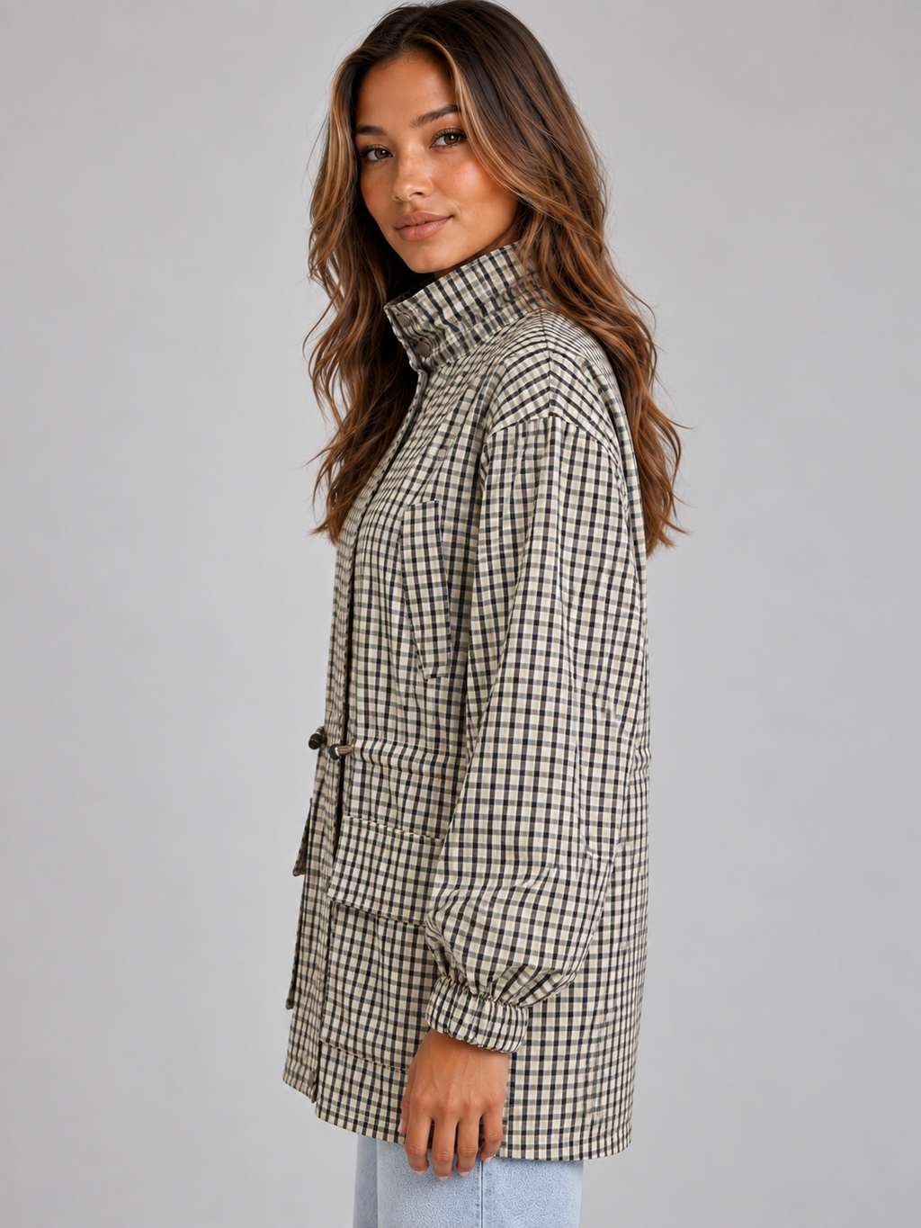 Woman wearing checkered black and white field coat with high neck and drawstring, styled with light blue jeans against gray background