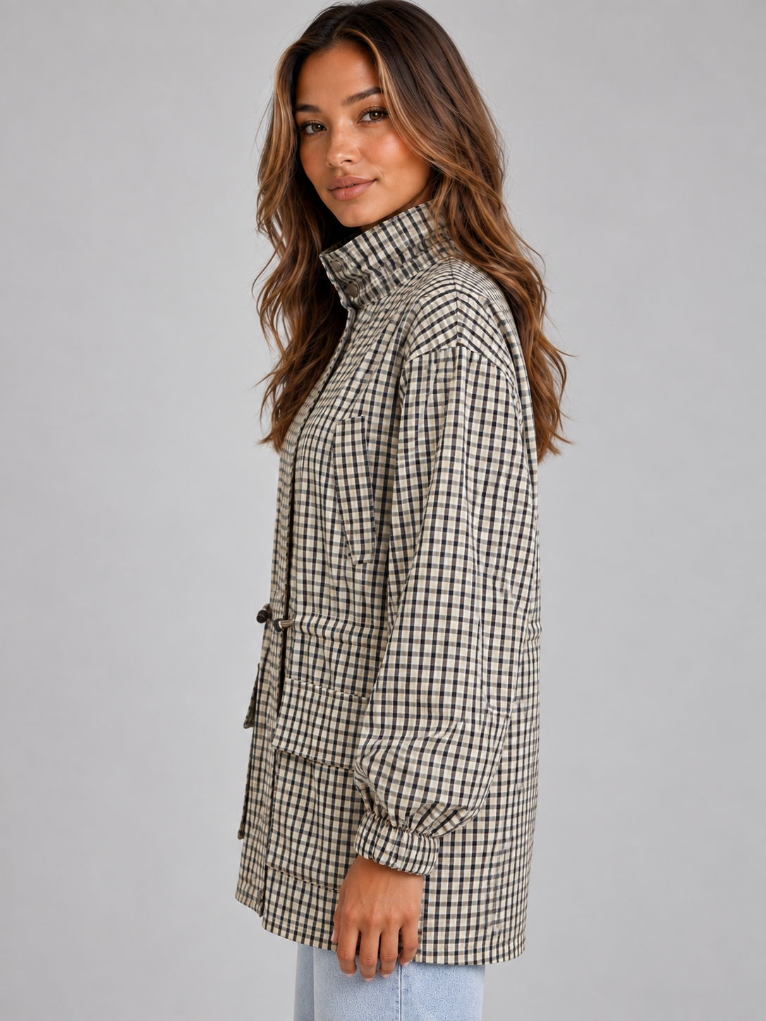 Woman wearing checkered black and white field coat with high neck and drawstring, styled with light blue jeans against gray background