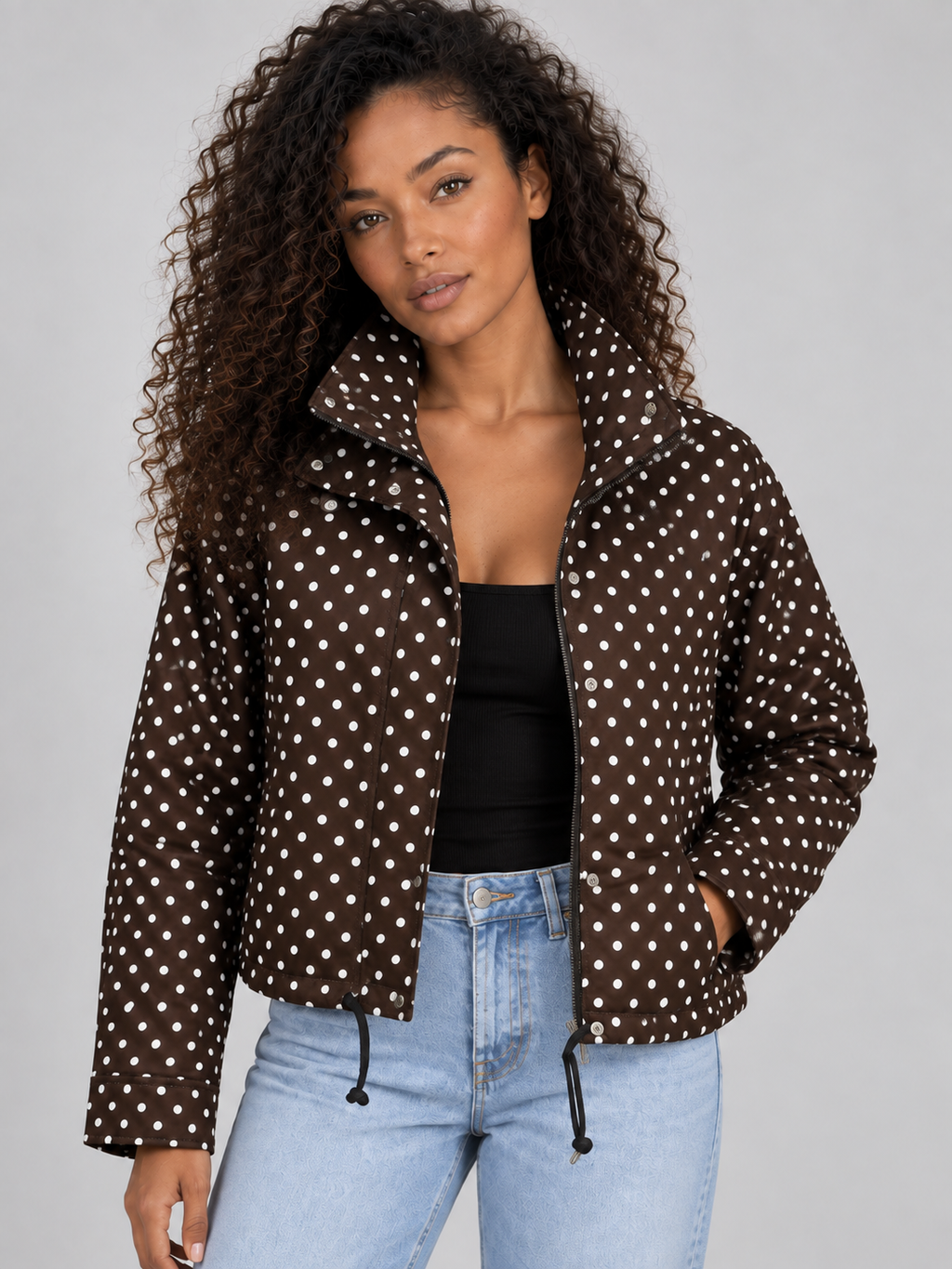 Woman wearing brown polka dot cropped zip-up jacket with high collar and drawstring, styled with black top and light blue jeans