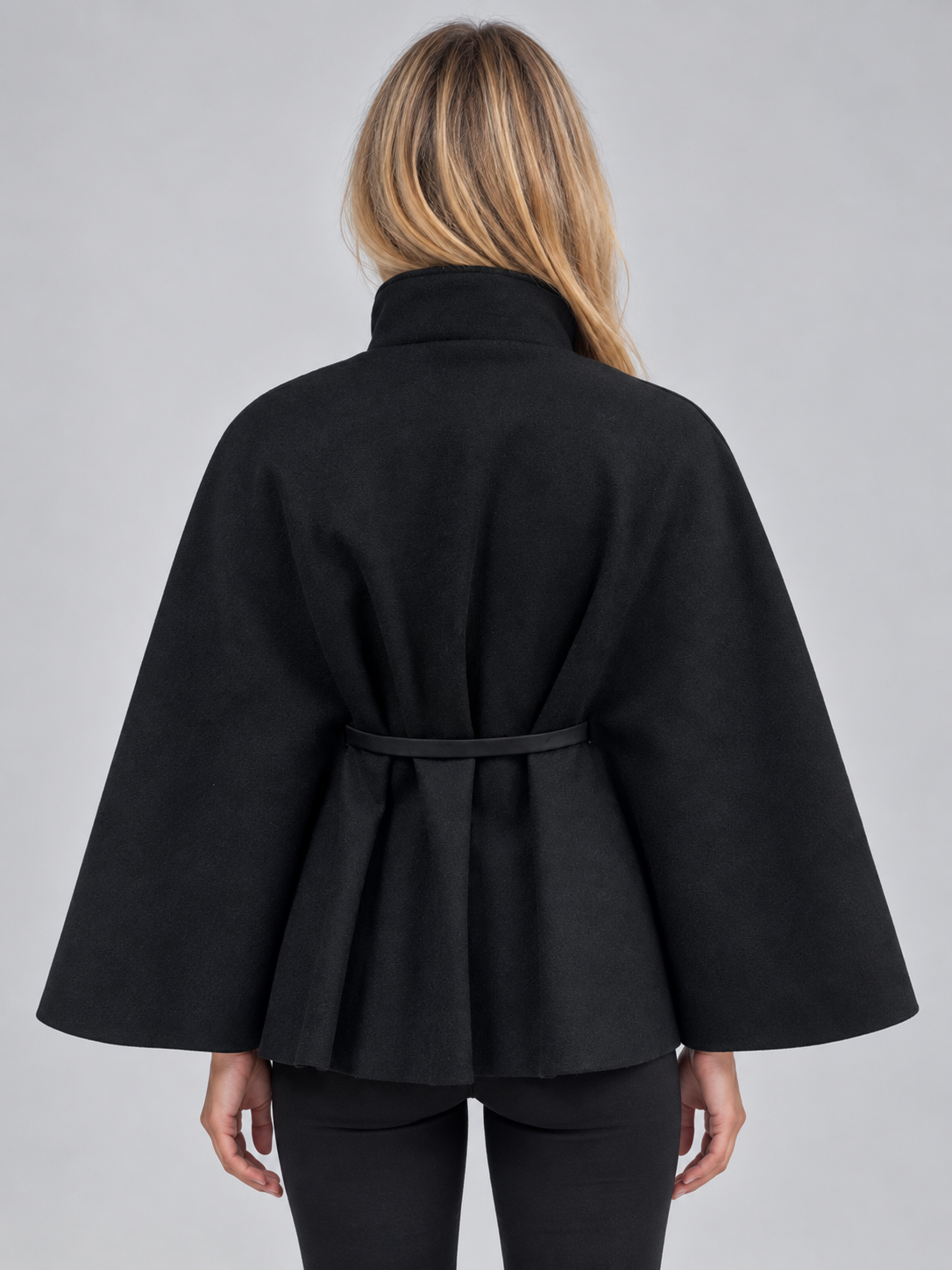 Woman wearing black belted cape jacket with flared sleeves and high neck, shown from back view against gray background