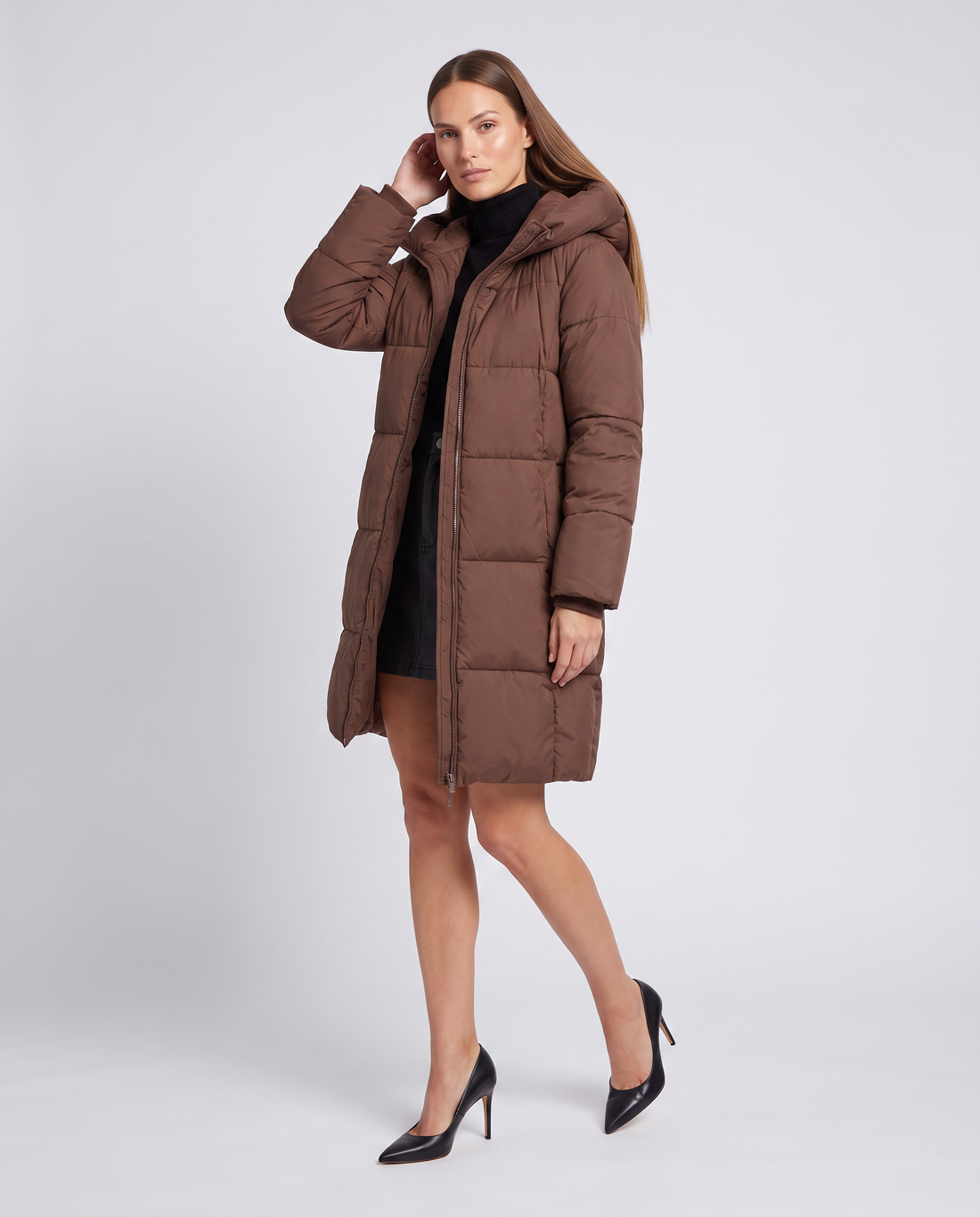 Woman wearing brown knee-length hooded puffer coat with black turtleneck and black heels against white background