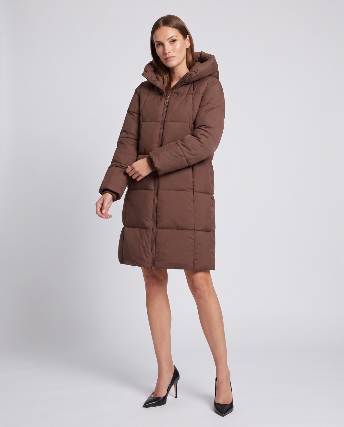 Woman wearing brown hooded knee-length puffer coat with zipper front and black heeled pumps on white background