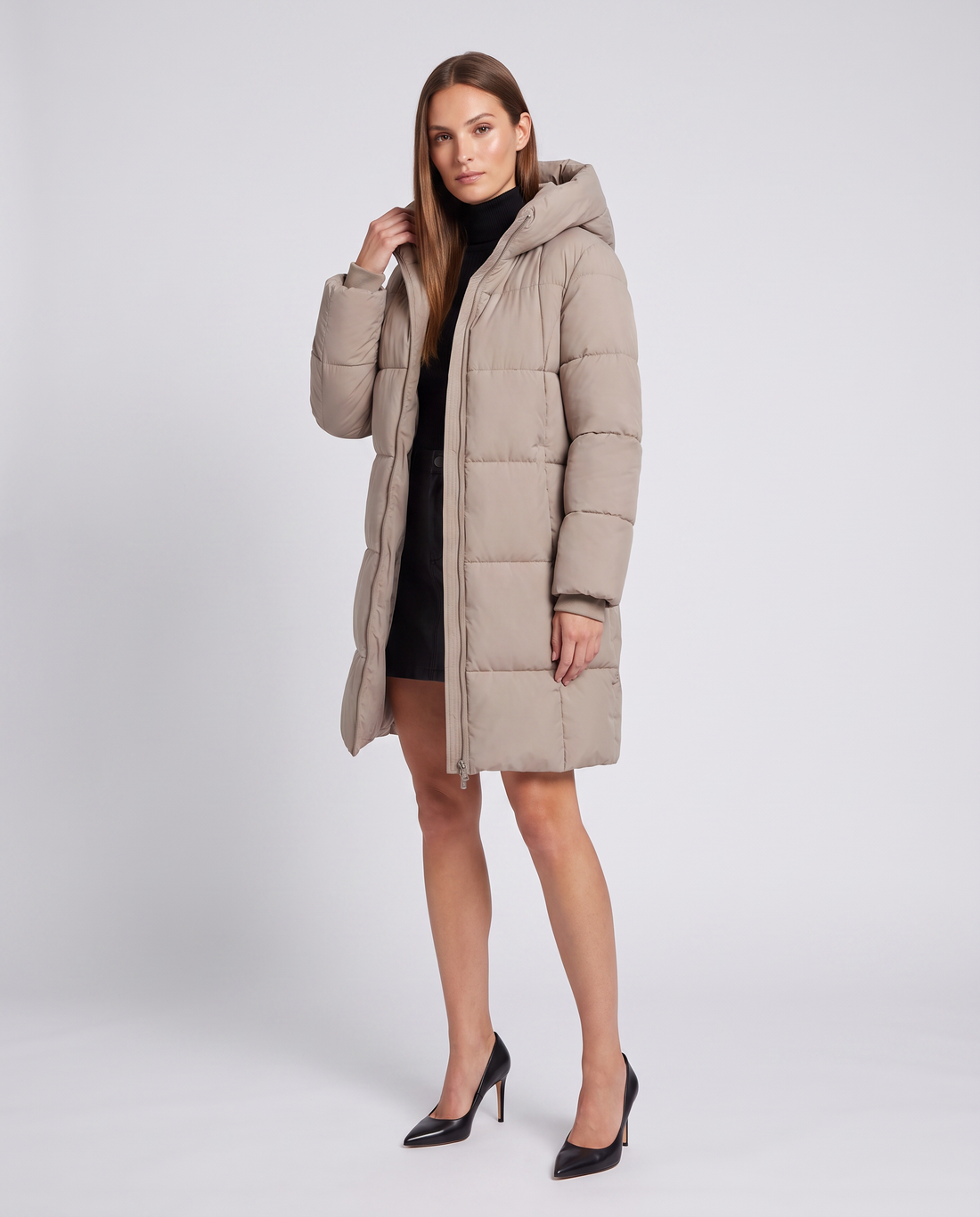 Woman wearing beige hooded puffer coat with black turtleneck and black heels against white background