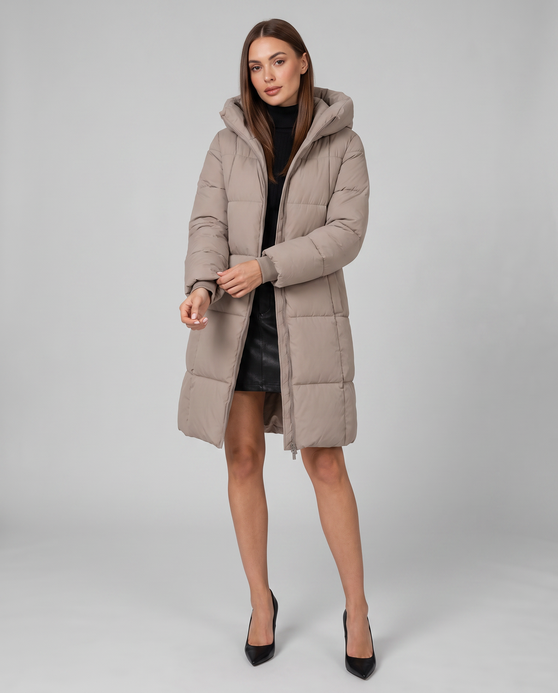 Woman wearing beige knee-length hooded puffer coat over black dress with black pointed-toe heels against gray background