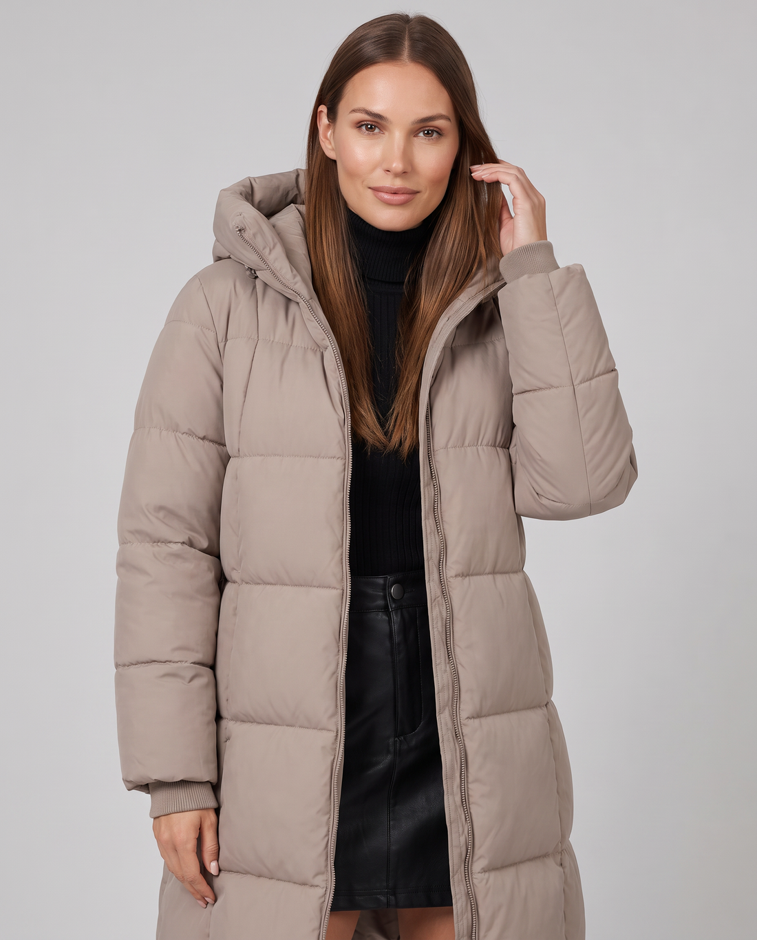 Woman wearing khaki knee-length hooded puffer coat with black turtleneck and black skirt, posed against gray background