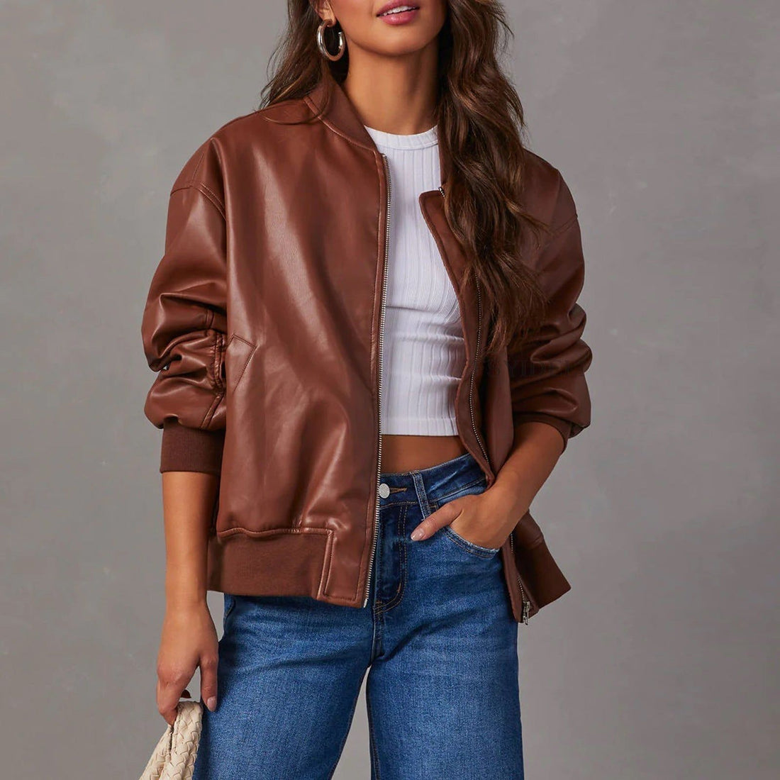 Woman wearing cognac brown leather bomber jacket with white ribbed top and blue jeans, holding cream woven handbag