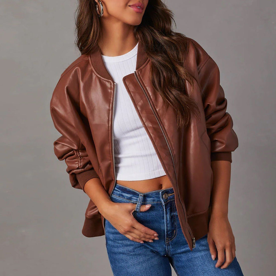 Woman wearing brown leather jacket over white ribbed crop top and blue jeans, silver hoop earrings, gray background