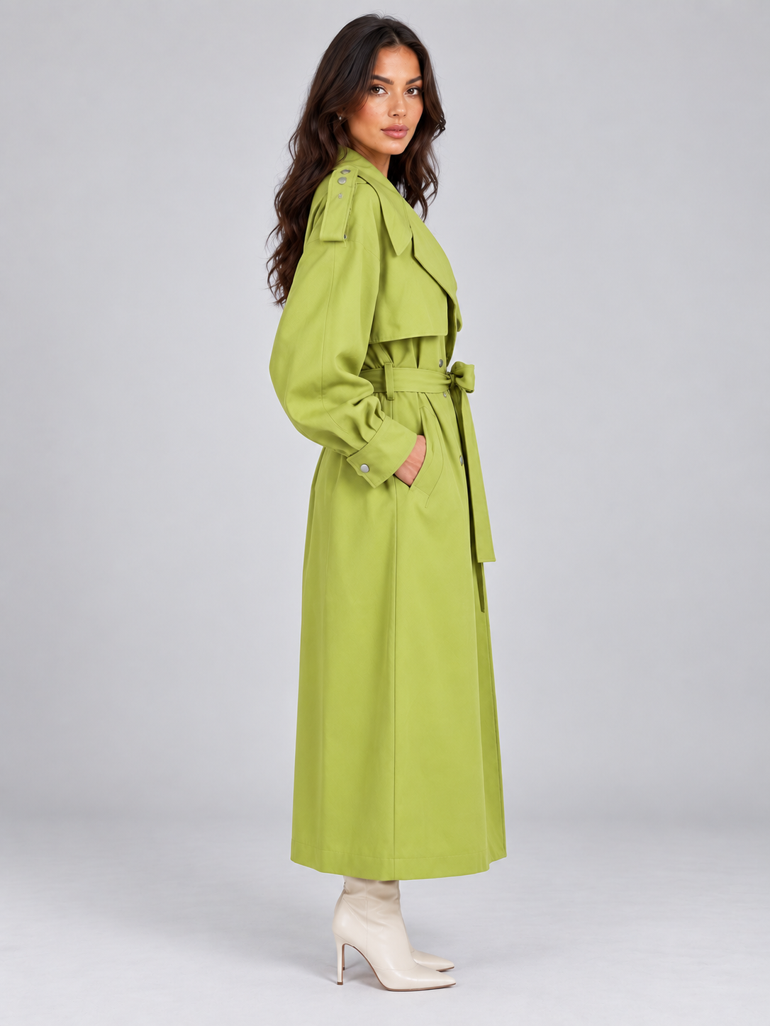 Woman wearing lime green double breasted belted trench coat with long sleeves and cream white heeled boots against gray background