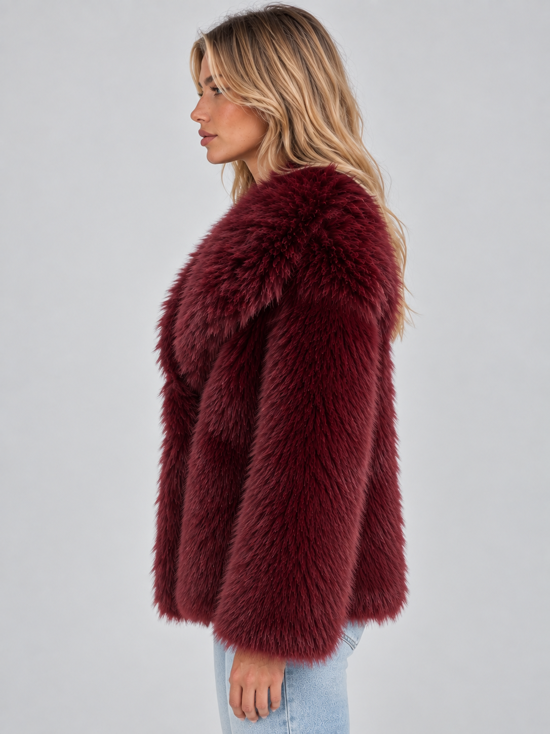 Woman wearing burgundy oversized faux fur winter coat with plush collar over light blue jeans, shown in profile view
