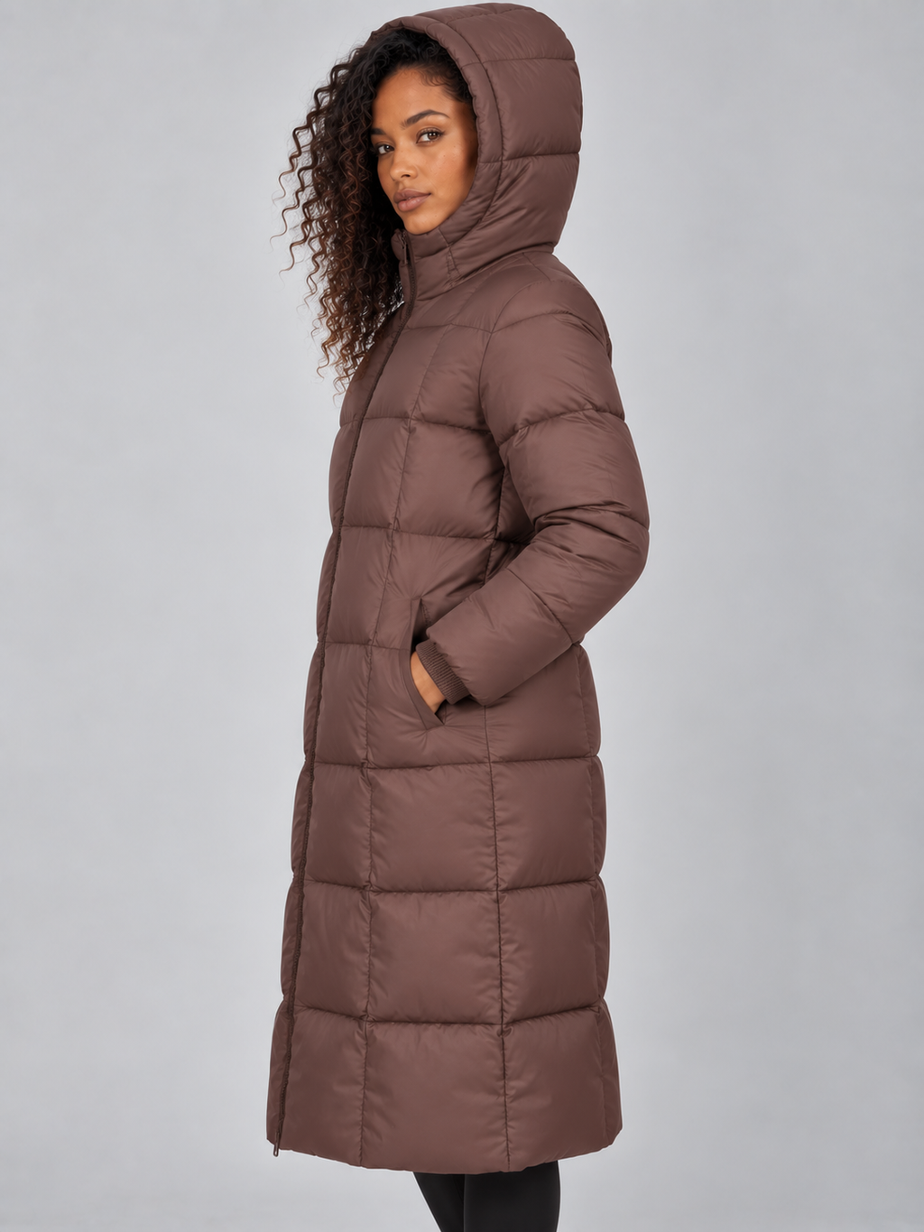 Woman wearing brown long hooded puffer jacket with hands in pockets, viewed from side angle against white background