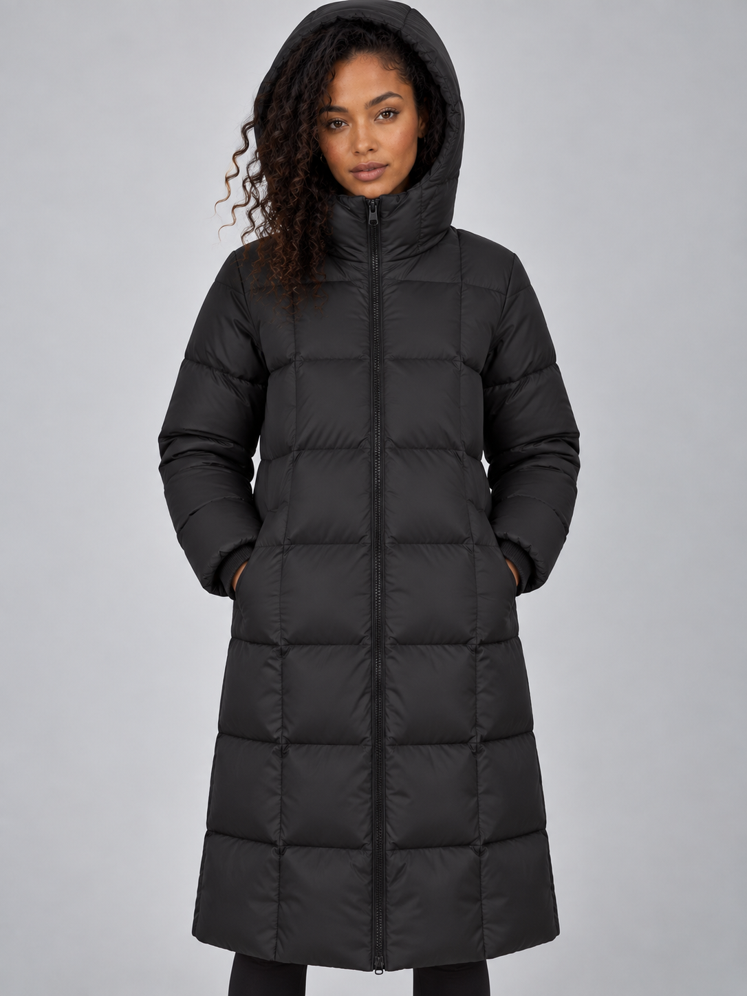 Black padded puffer jacket with hood, full zip front, and long length worn by woman against gray background