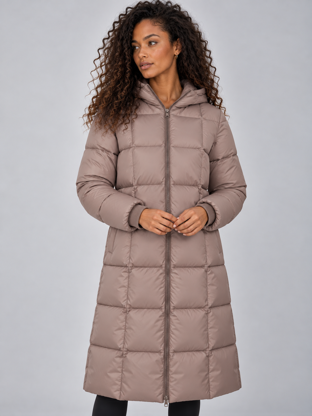 Woman wearing taupe padded puffer jacket with hood, full zip, and long length over black pants
