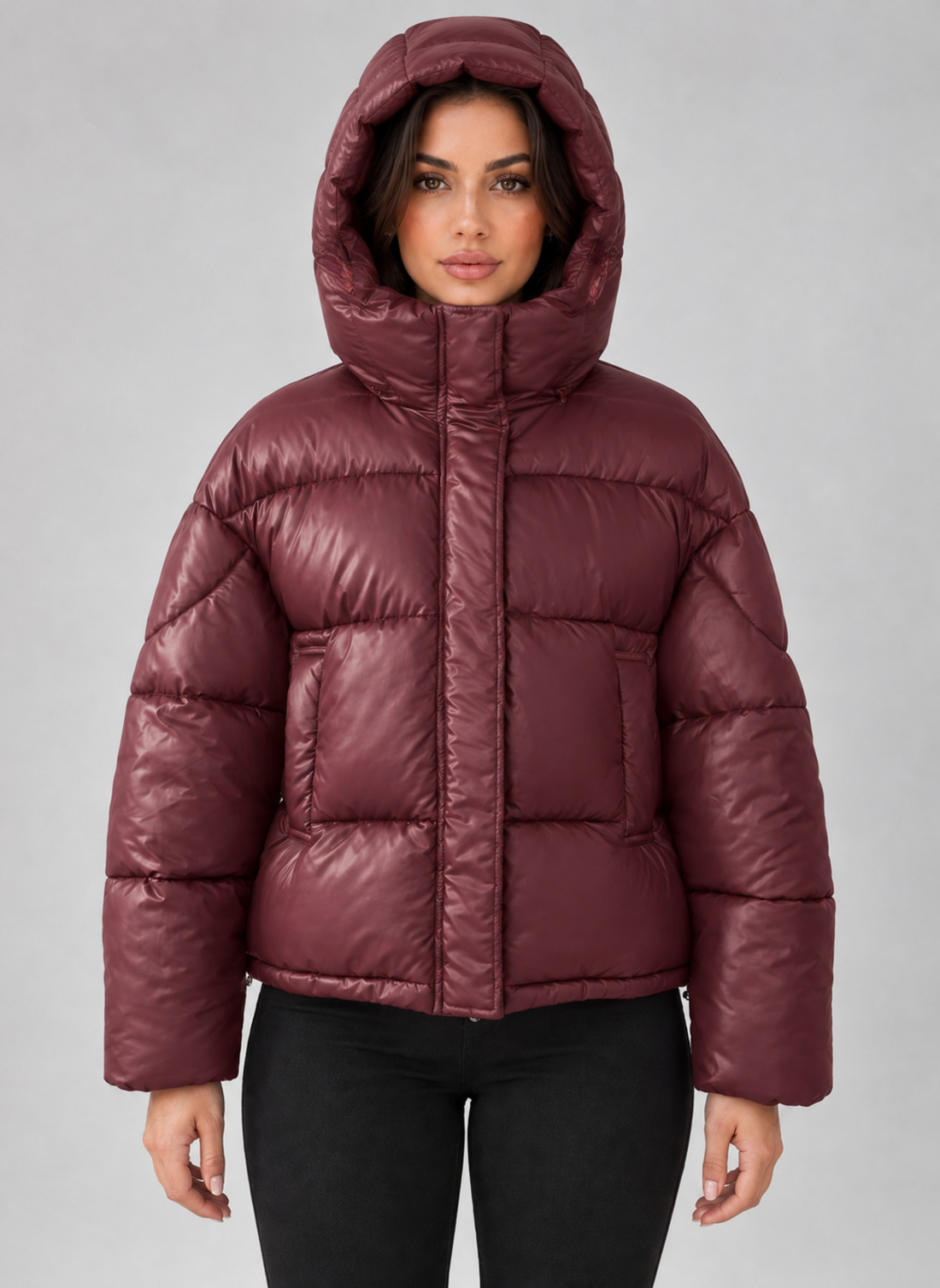 Woman wearing burgundy padded puffer jacket with hood and full zip, paired with black pants against gray background