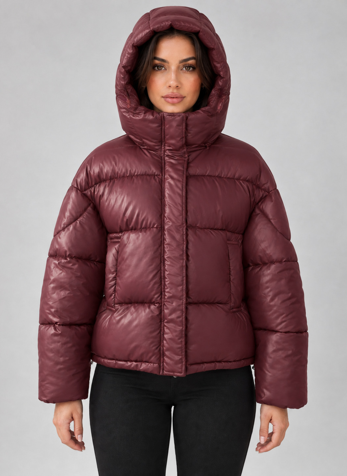 Woman wearing burgundy padded puffer jacket with hood and full zip, paired with black pants against gray background