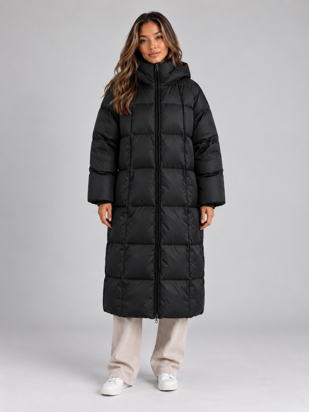 Woman wearing black padded long hooded puffer coat with full zip, beige pants, and white sneakers