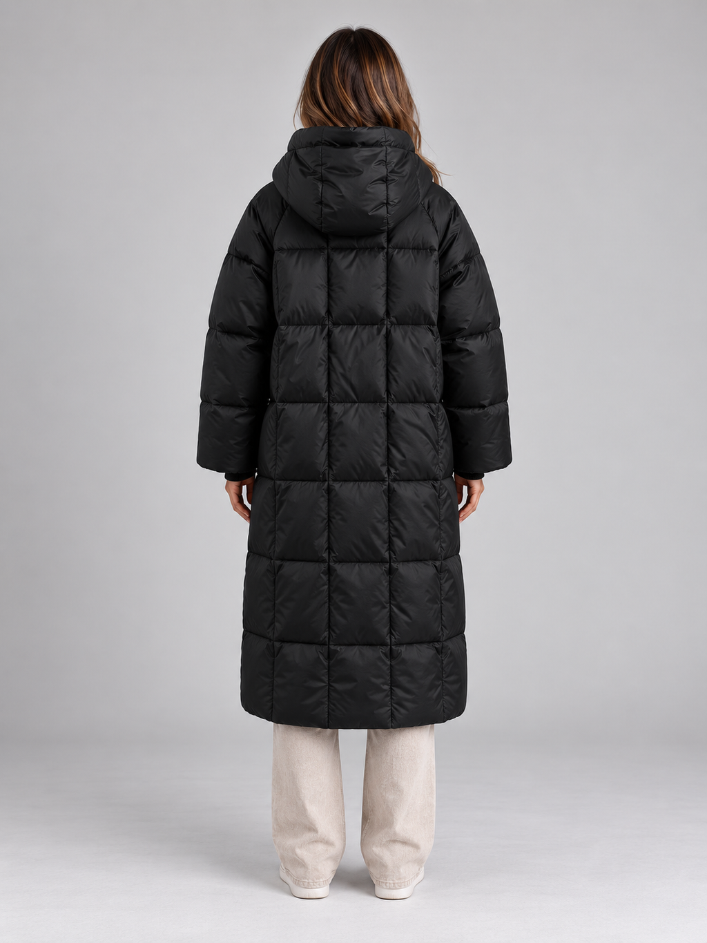 Woman wearing black padded long hooded puffer coat with full zip over cream pants, back view