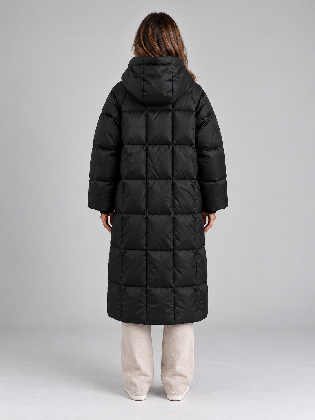 Woman wearing black padded long hooded puffer coat with full zip over cream pants, back view