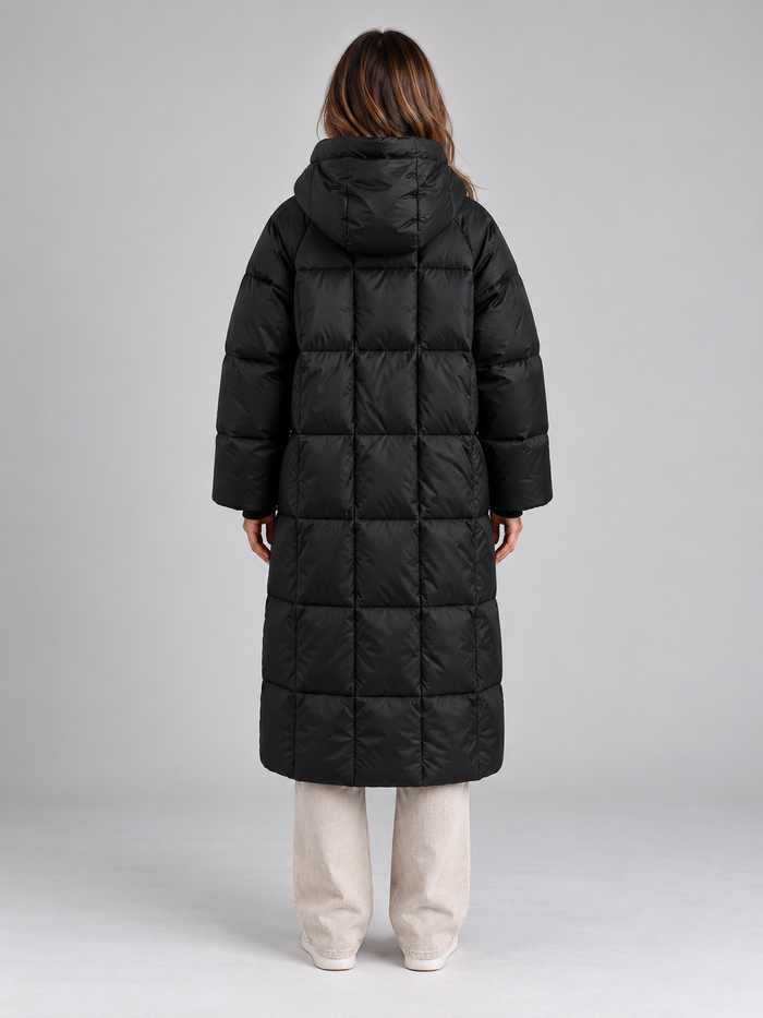 Woman wearing black padded long hooded puffer coat with full zip over cream pants, back view