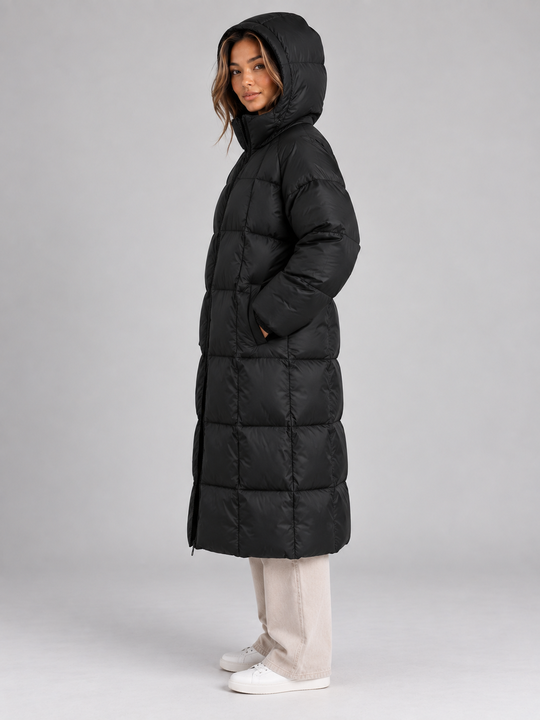 Woman wearing black padded long hooded puffer coat with full zip, styled with cream jeans and white sneakers