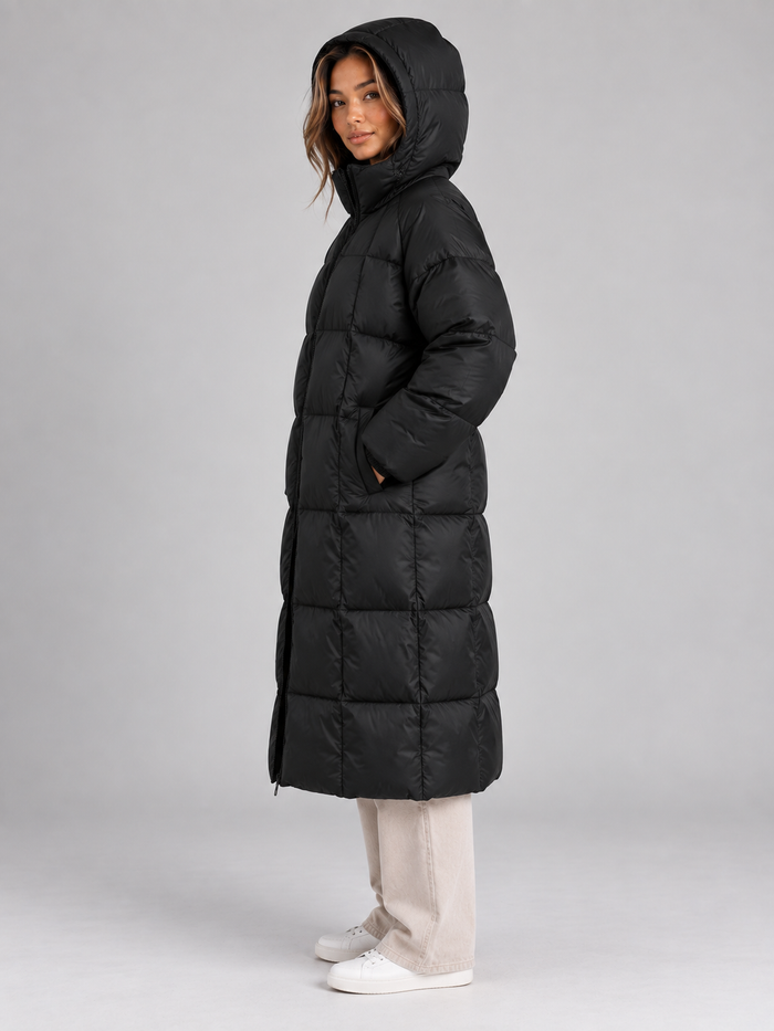 Woman wearing black padded long hooded puffer coat with full zip, styled with cream jeans and white sneakers