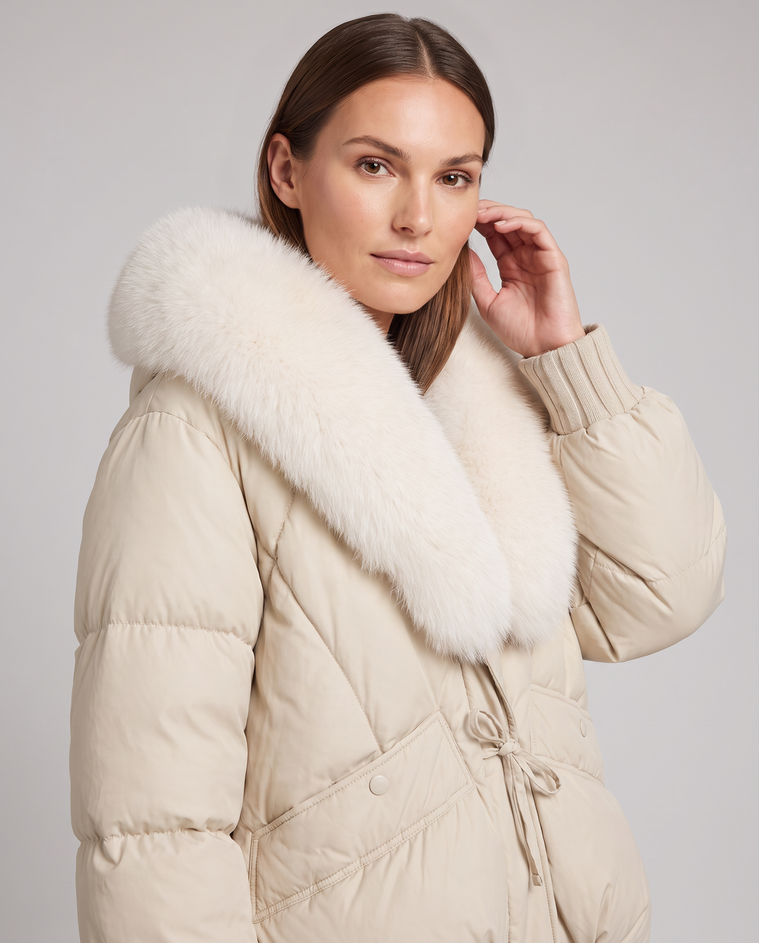 Woman wearing beige padded puffer jacket with white fur collar, ribbed cuffs, and drawstring waist against gray background