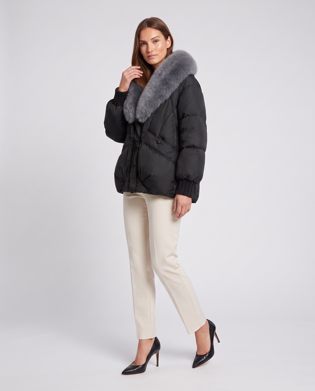 Woman wearing black padded puffer jacket with gray fur collar, cream pants, and black pointed-toe heels on white background