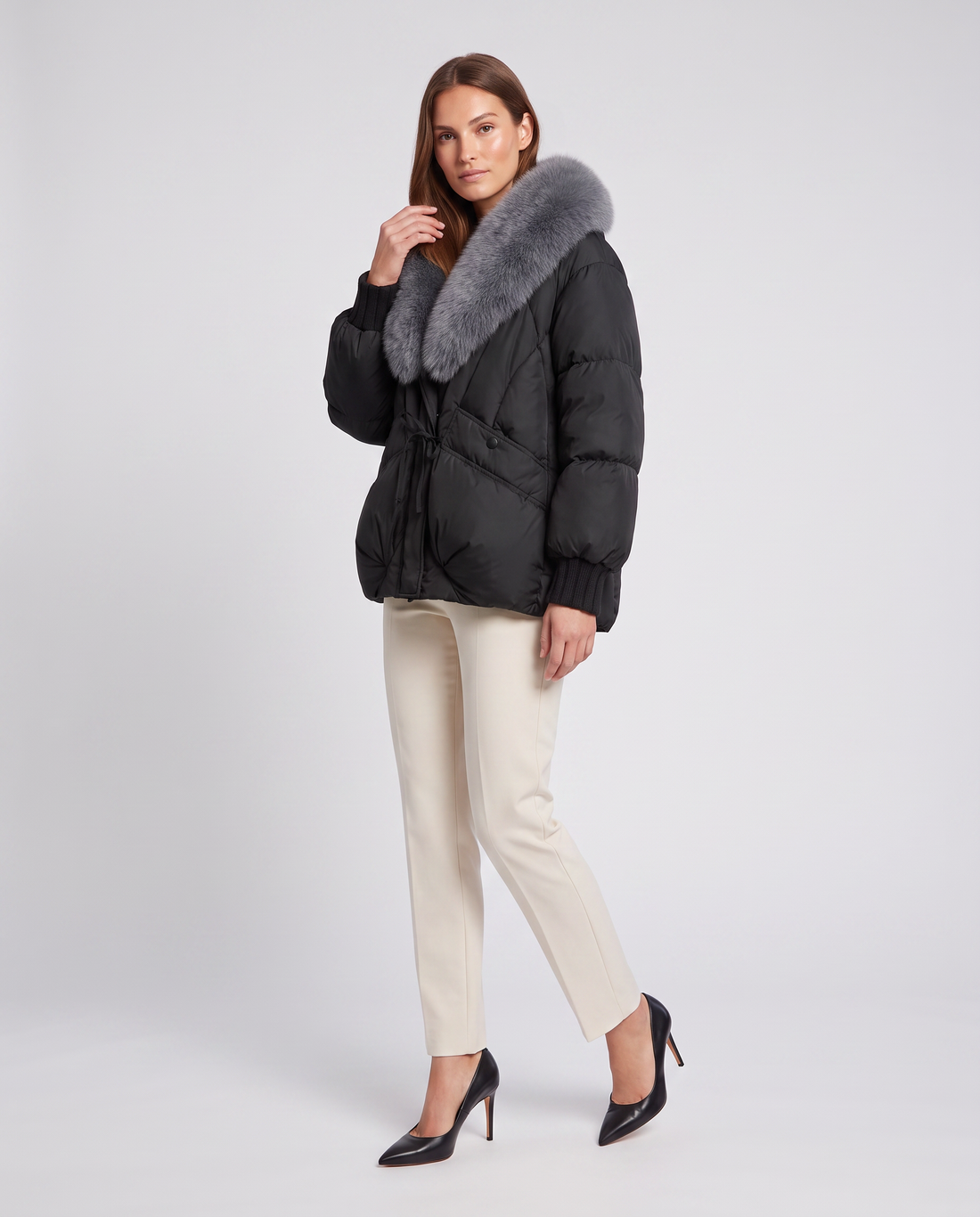 Woman wearing black padded puffer jacket with gray fur collar, cream pants, and black pointed-toe heels on white background