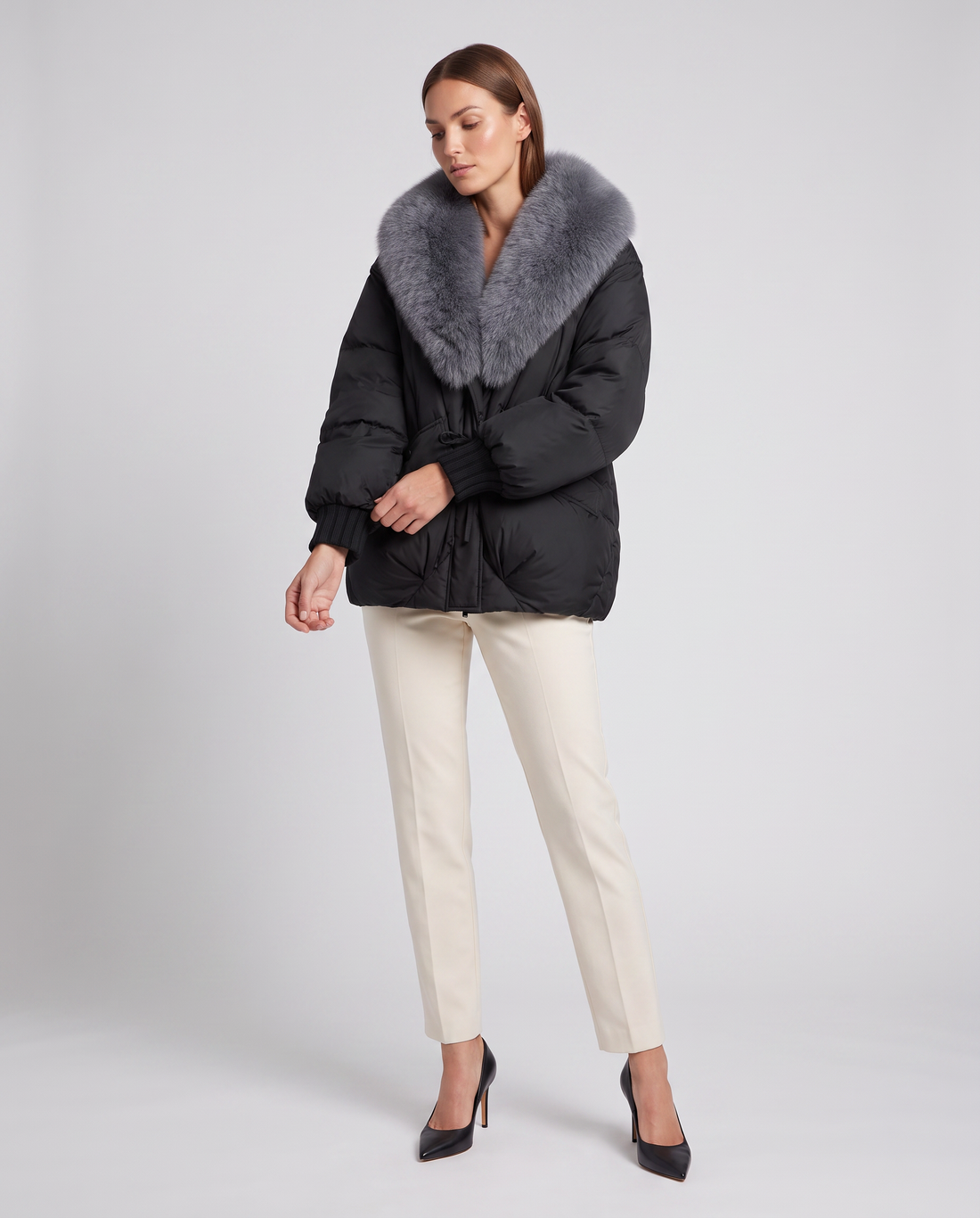 Woman wearing black padded puffer jacket with gray fur collar, cream pants, and black heels