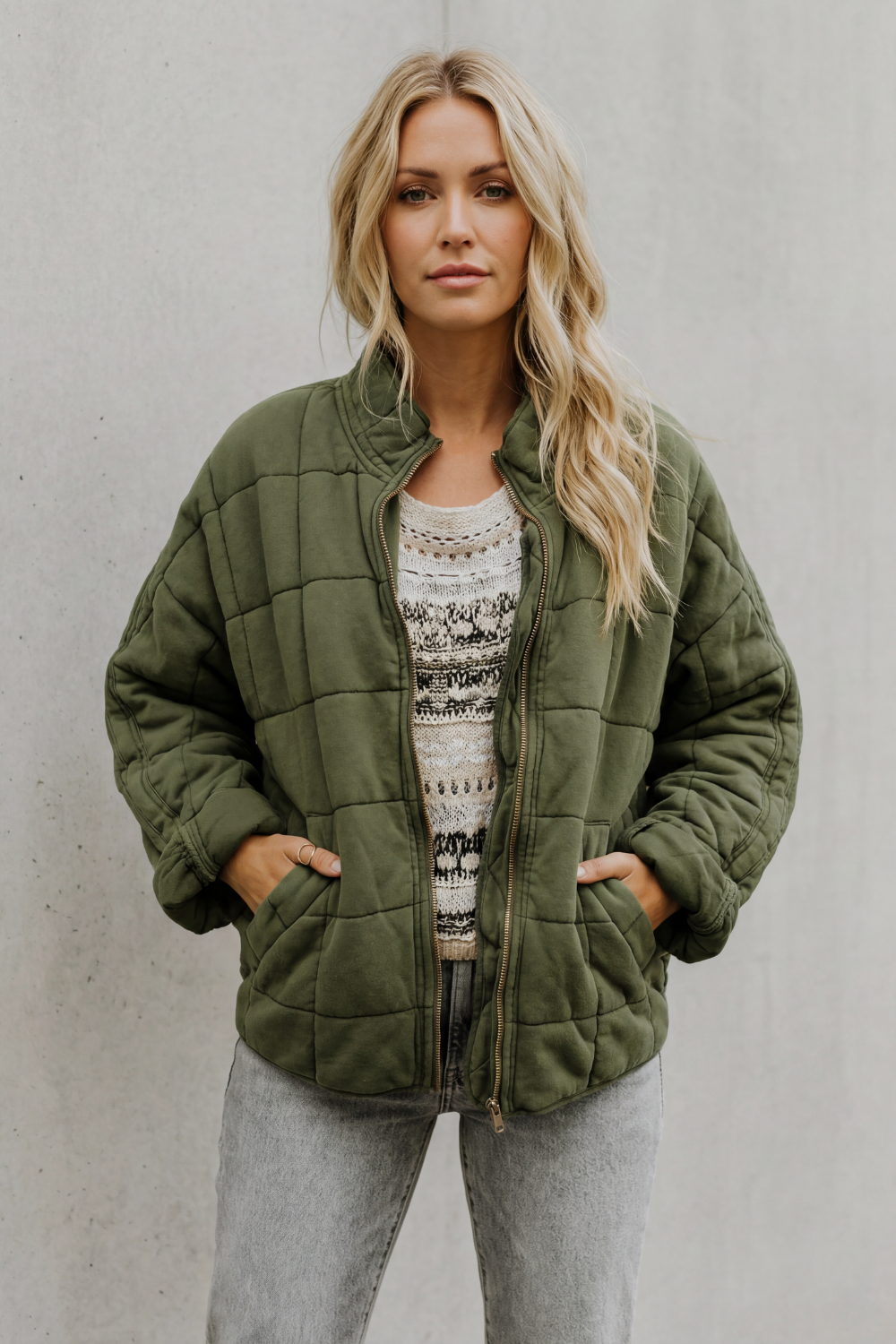 Woman wearing olive green quilted jacket with high collar over patterned sweater and light gray jeans