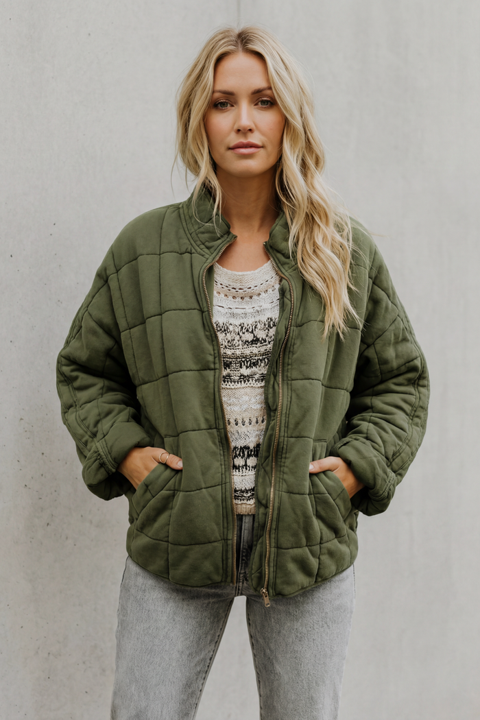 Woman wearing olive green quilted jacket with high collar over patterned sweater and light gray jeans