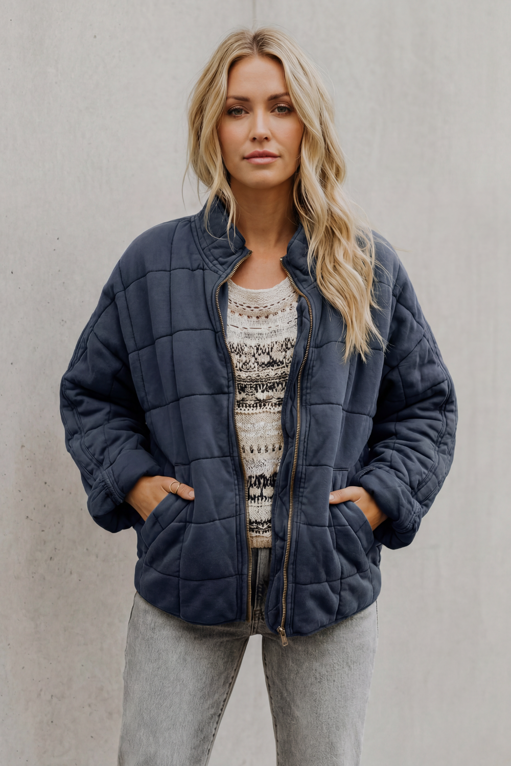 Woman wearing navy quilted jacket with high collar over patterned sweater and gray pants, hands in pockets against white background