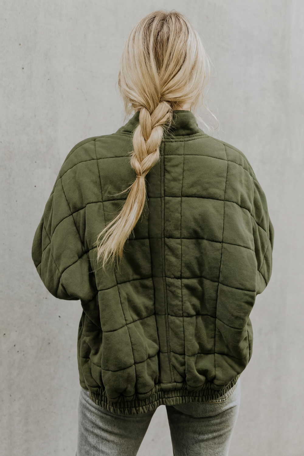 Woman wearing sage green quilted jacket with high collar and braided blonde hair, back view
