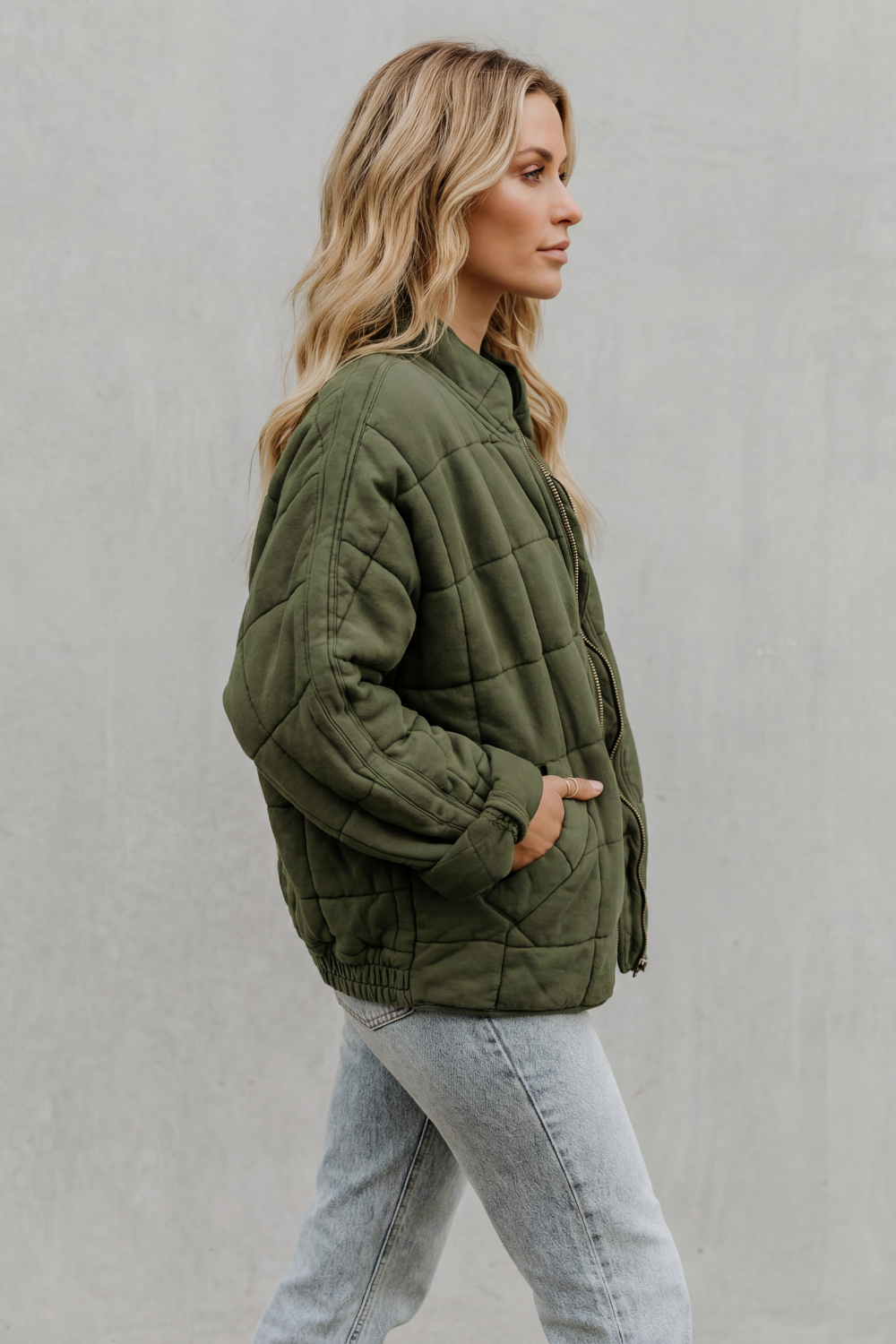Woman wearing olive green quilted jacket with high collar and light wash jeans, shown from side profile against neutral background