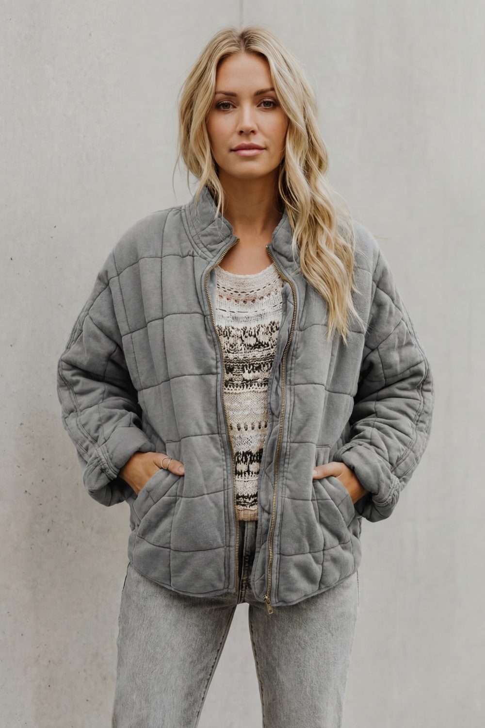 Woman wearing gray quilted jacket with high collar over beige patterned sweater and gray jeans, hands in pockets