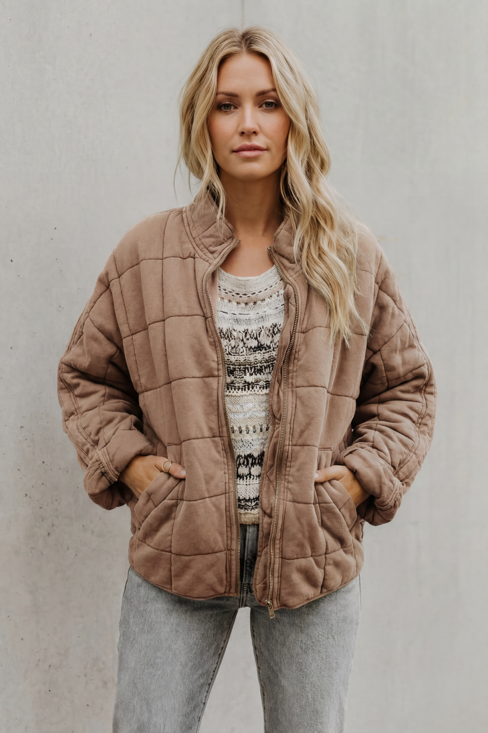 Woman wearing tan quilted jacket with high collar over patterned sweater and gray jeans, standing against white background