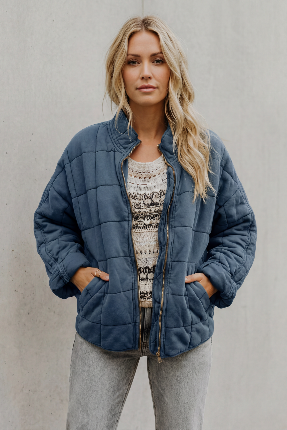 Woman wearing navy blue quilted jacket with high collar over beige patterned sweater and gray pants against white background