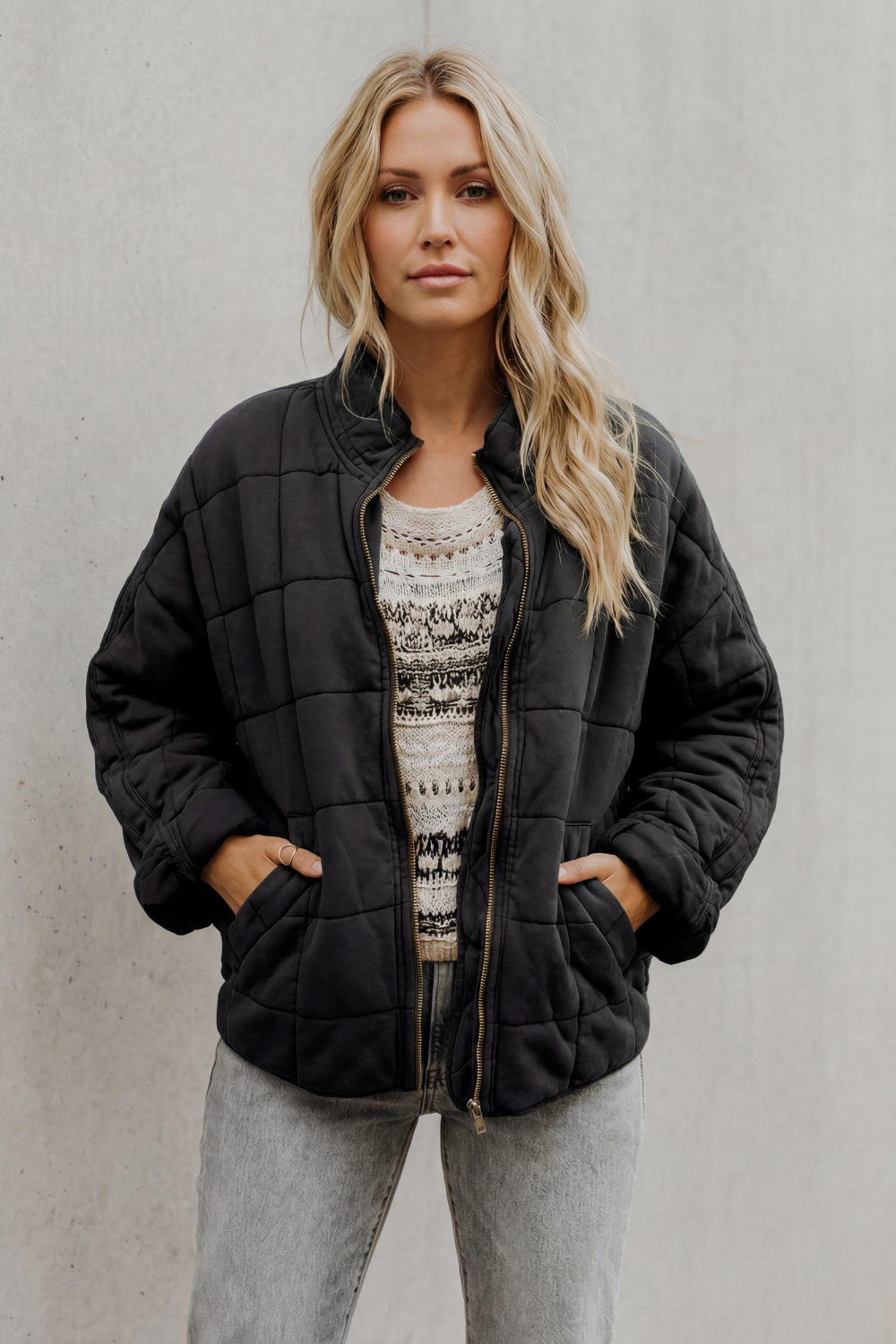 Woman wearing black quilted jacket with high collar, geometric patterned sweater, and light gray pants against white background