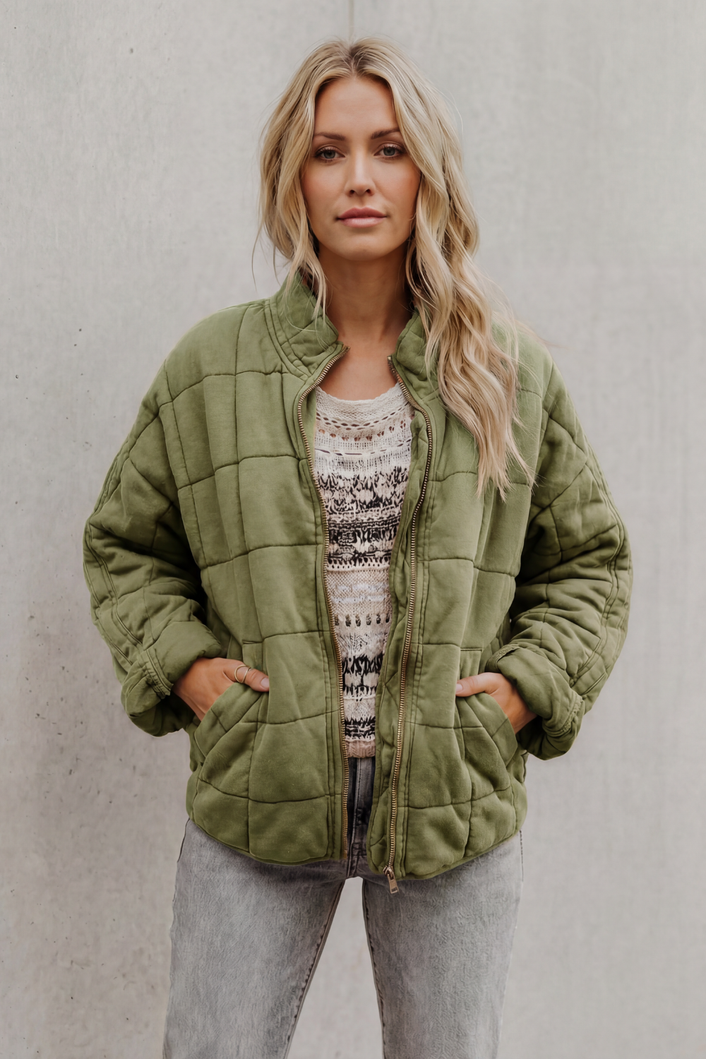 Woman wearing olive green quilted jacket with high collar and zip front over patterned sweater and gray pants