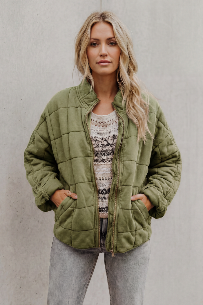 Woman wearing olive green quilted jacket with high collar and zip front over patterned sweater and gray pants