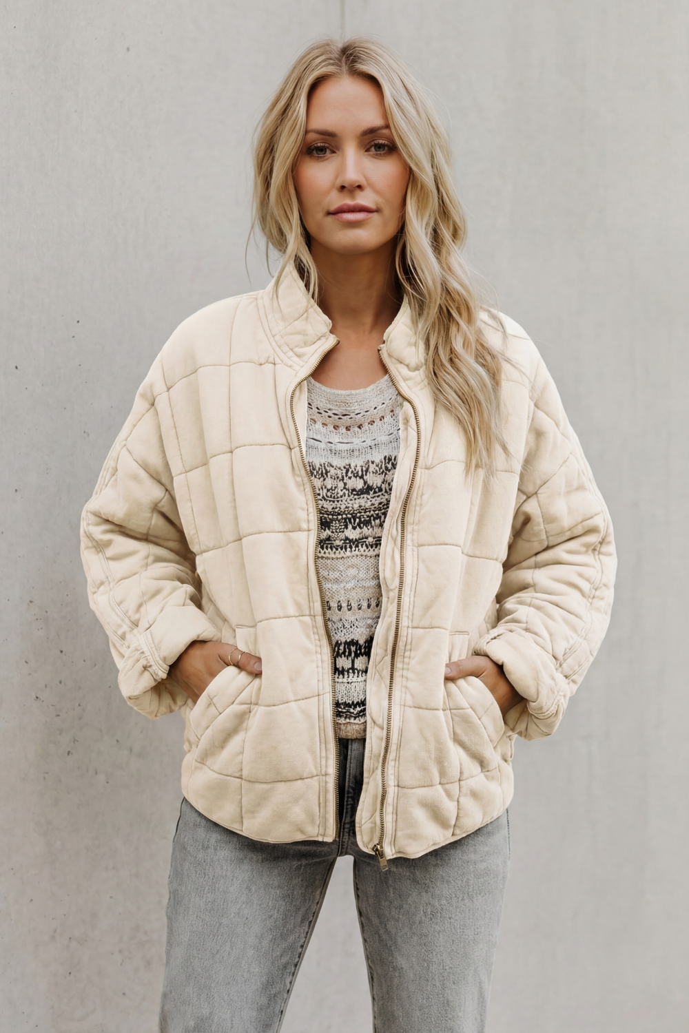 Woman wearing cream quilted jacket with high collar over patterned sweater and gray jeans, hands in pockets