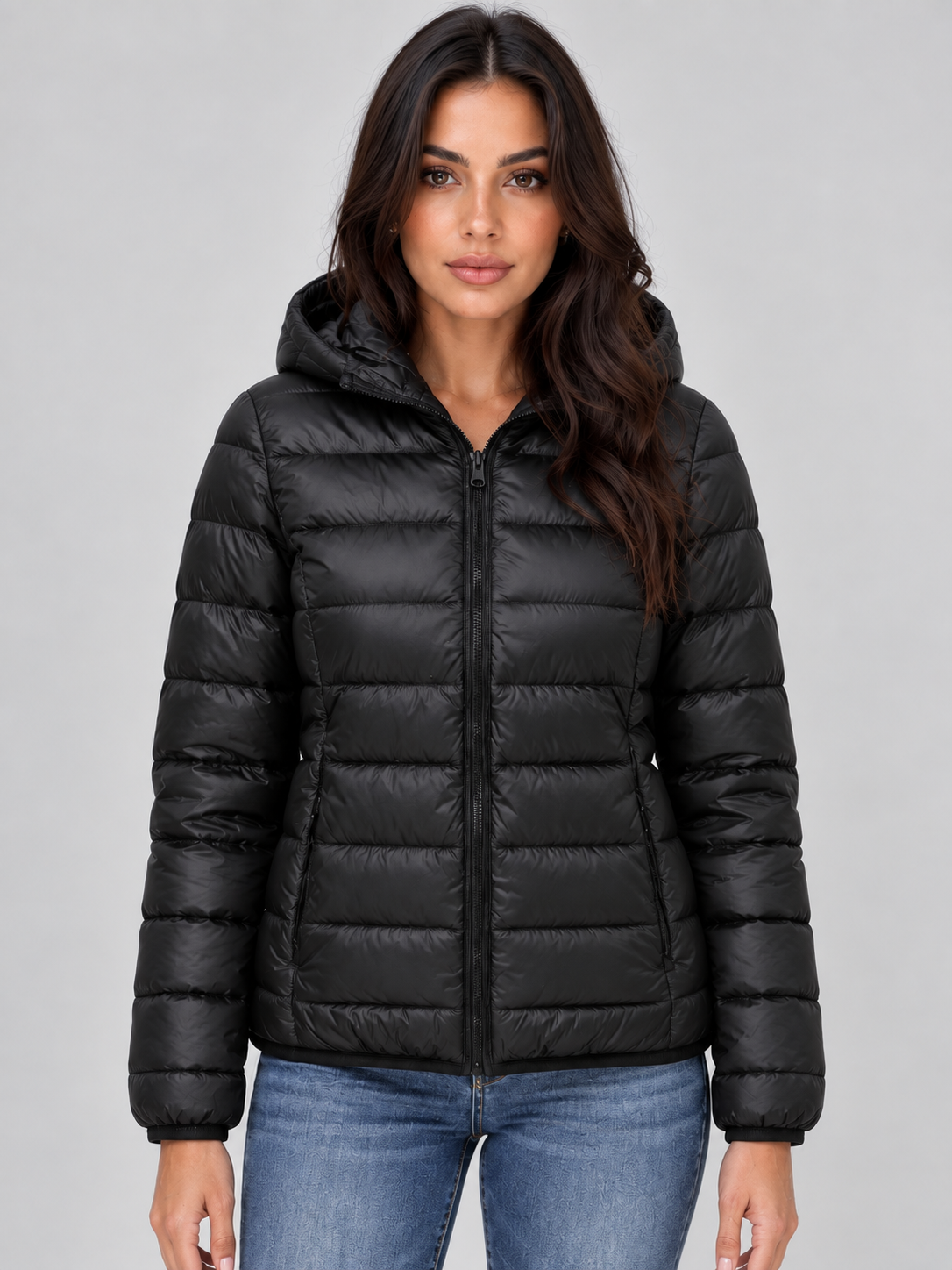 Woman wearing black quilted zip-up hooded puffer jacket with horizontal stitching, styled with blue jeans against gray background