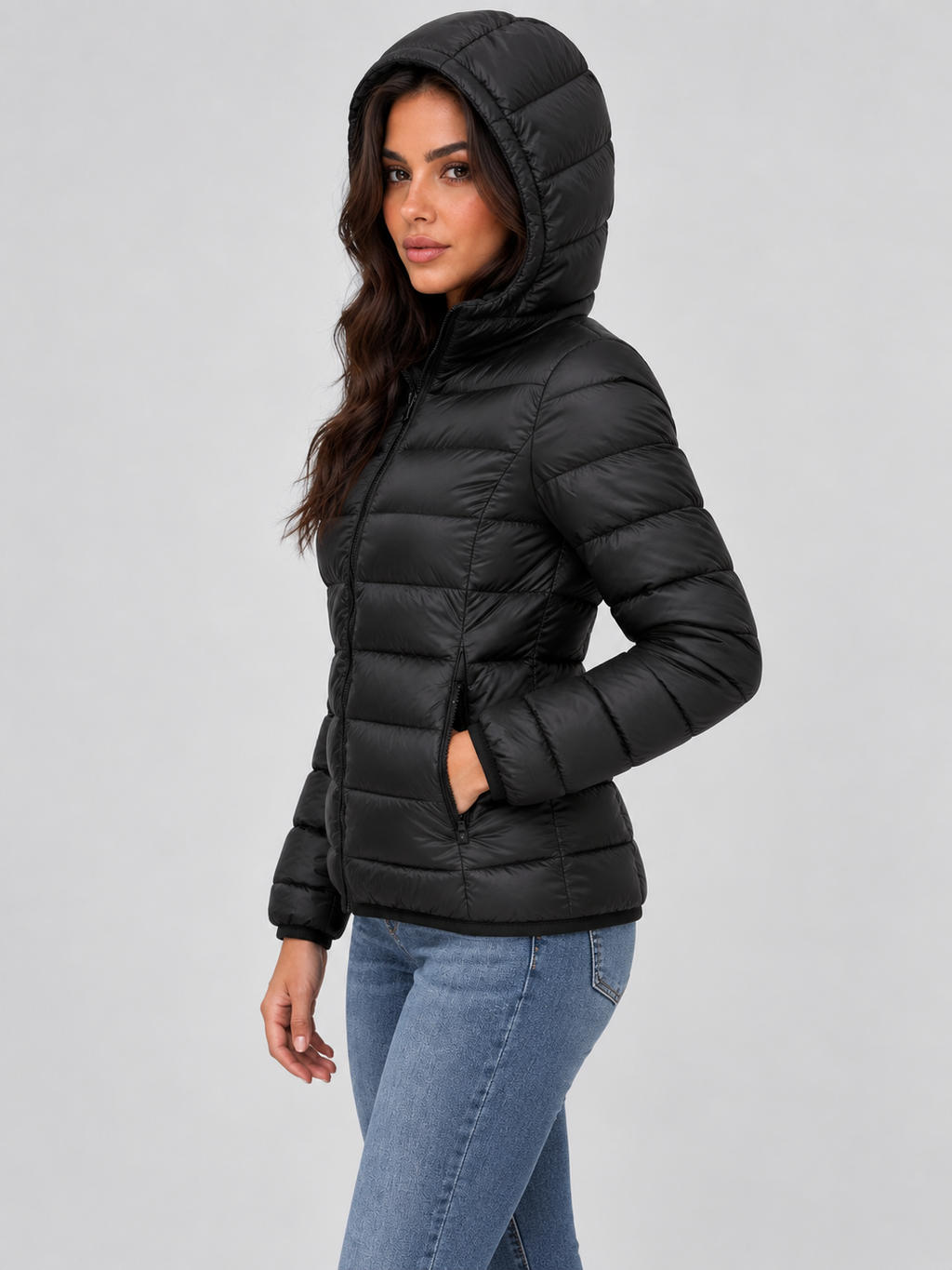 Woman wearing black quilted puffer jacket with hood and front zip, paired with blue jeans against gray background