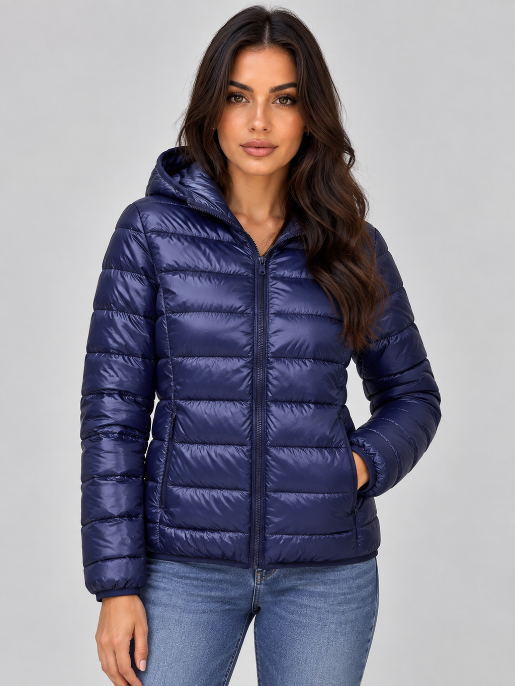 Woman wearing navy blue quilted zip-up hooded puffer jacket with jeans against white background
