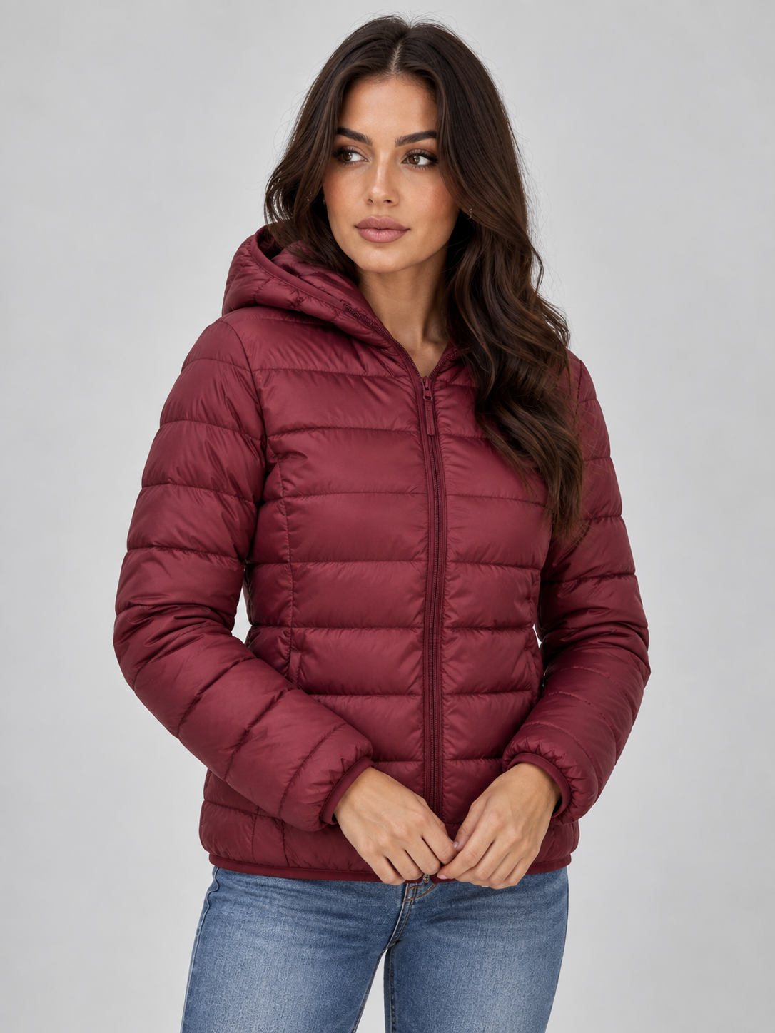Woman wearing burgundy quilted zip-up hooded puffer jacket over blue jeans, standing against white background