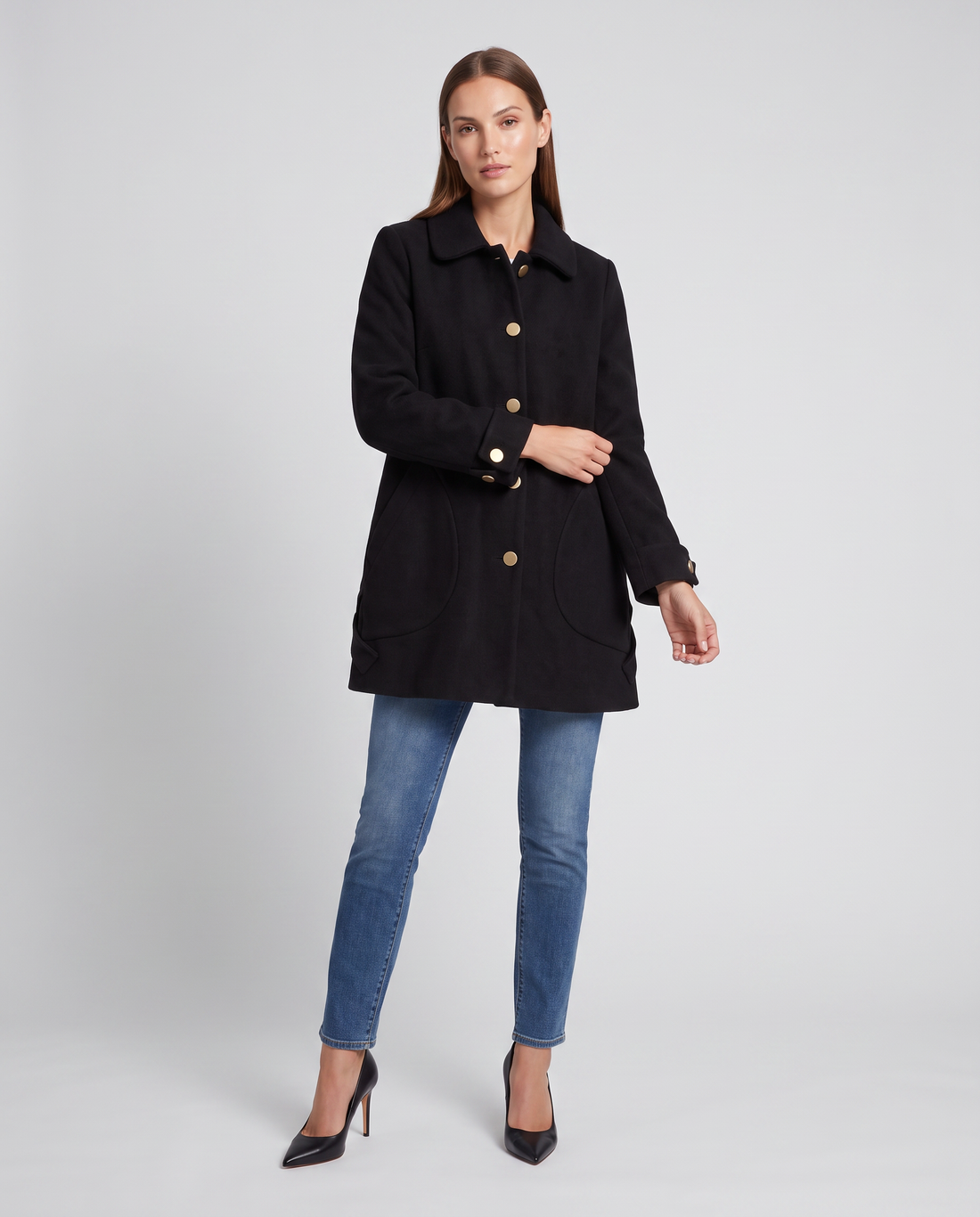 Woman wearing black round collar mid-length oversized coat with gold buttons over blue jeans and black heels