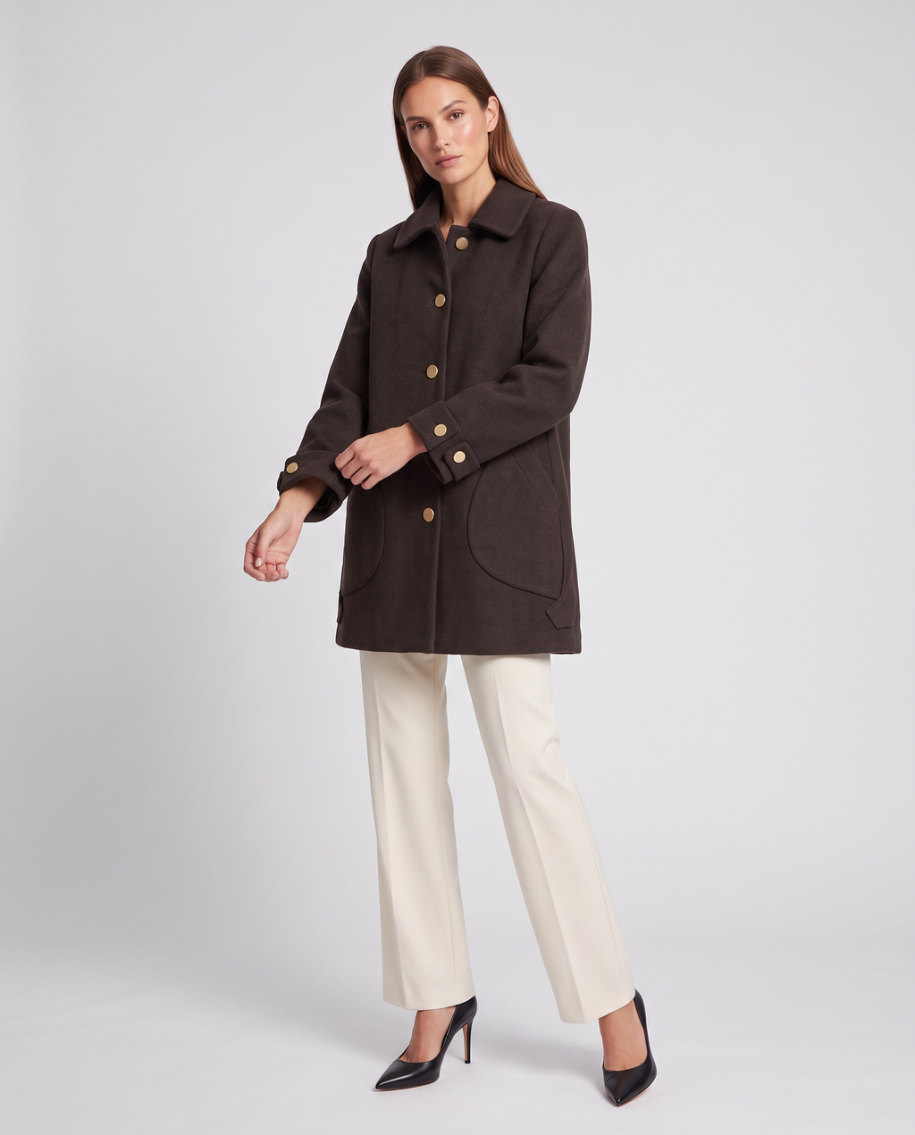 Woman wearing dark brown oversized coat with round collar and gold buttons over cream trousers and black heels
