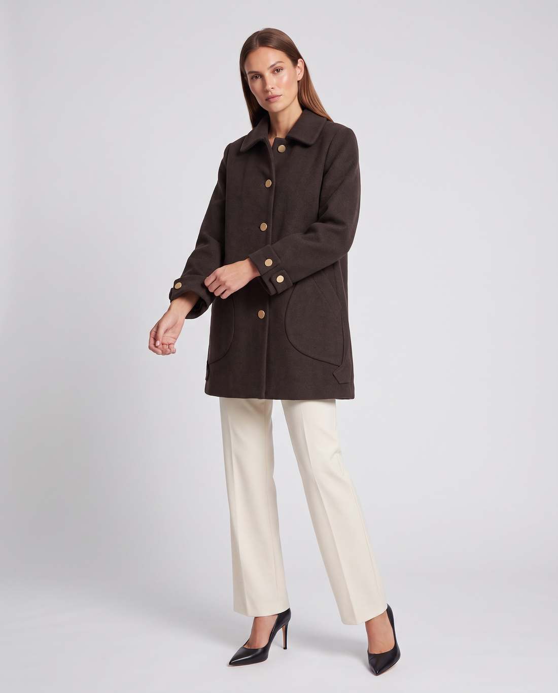 Woman wearing dark brown oversized coat with round collar and gold buttons over cream trousers and black heels