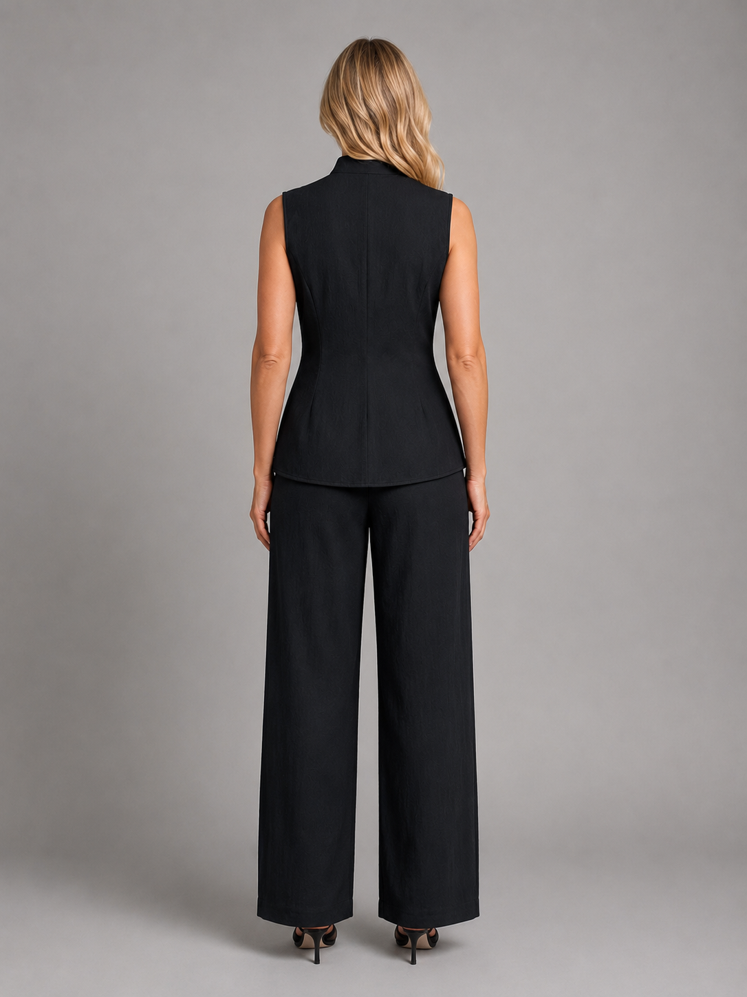 Woman wearing black sleeveless button-up vest top and wide-leg trousers, viewed from back, against neutral gray background